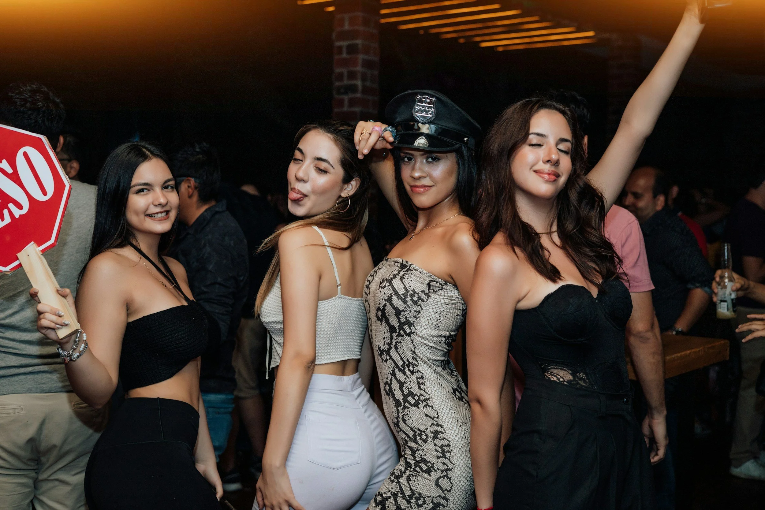 Four young women at a party, posing closely together with smiles and playful expressions, in a dimly lit setting with other people in the background.