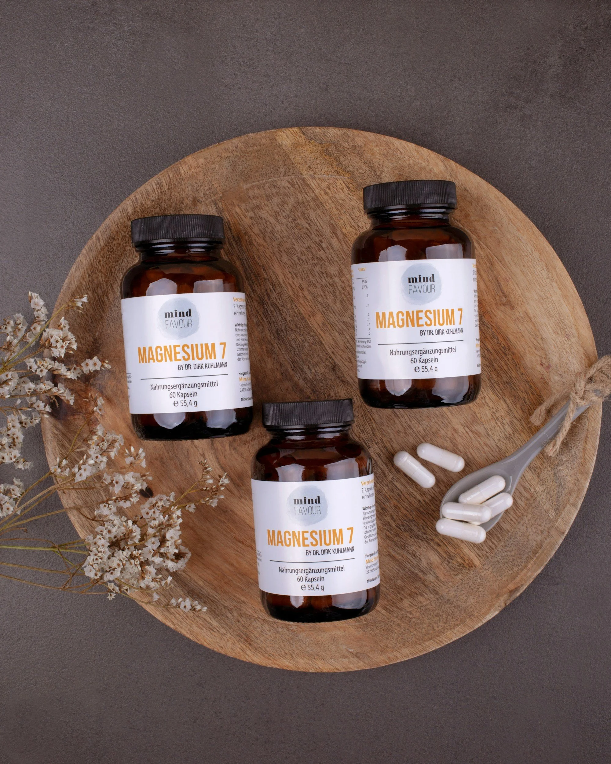 Three bottles of magnesium supplement labeled "Magnesium 7" by Dr. Dirk Kuhlmann on a round wooden tray, with a gray spoon holding white capsules and some capsules outside the bottles, against a gray background. Companies may wish to add seaweed.