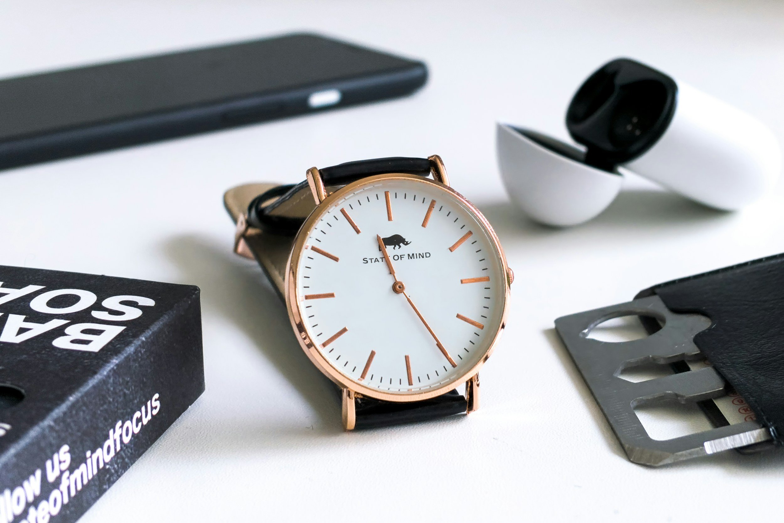 Close-up of a wristwatch with a white dial and rose gold casing, on a table with a smartphone, earbud case, and a multitool in a leather holder.