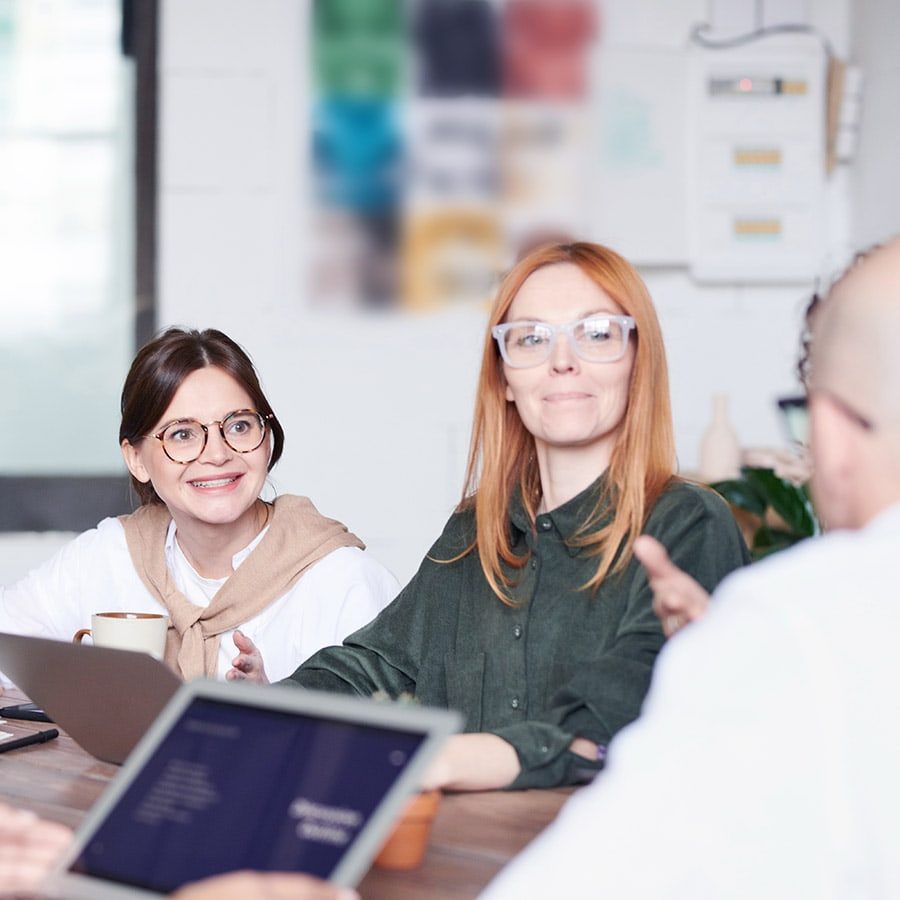 Group of people having a meeting, two women with glasses smiling and talking, laptops on the table, blurred background.