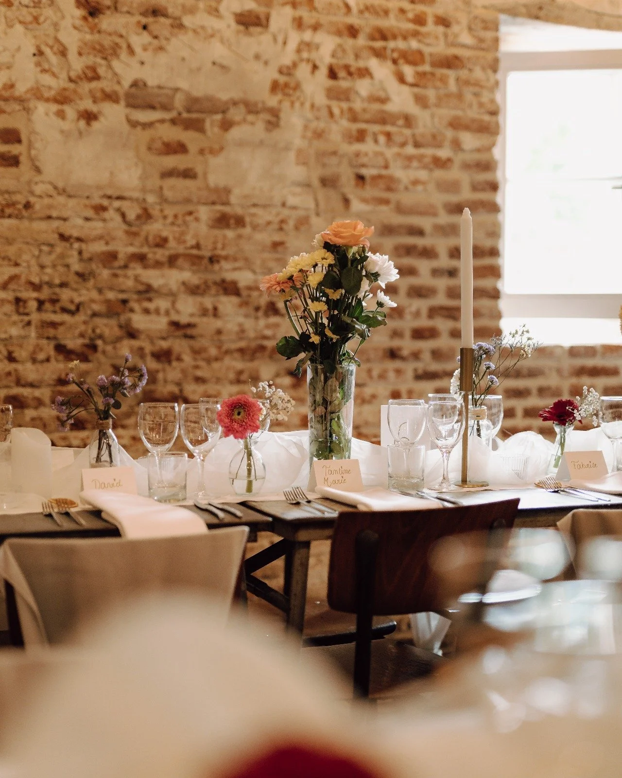 A wedding or special event table decorated with flowers, candles, and glassware against an exposed brick wall, with a window providing natural light.