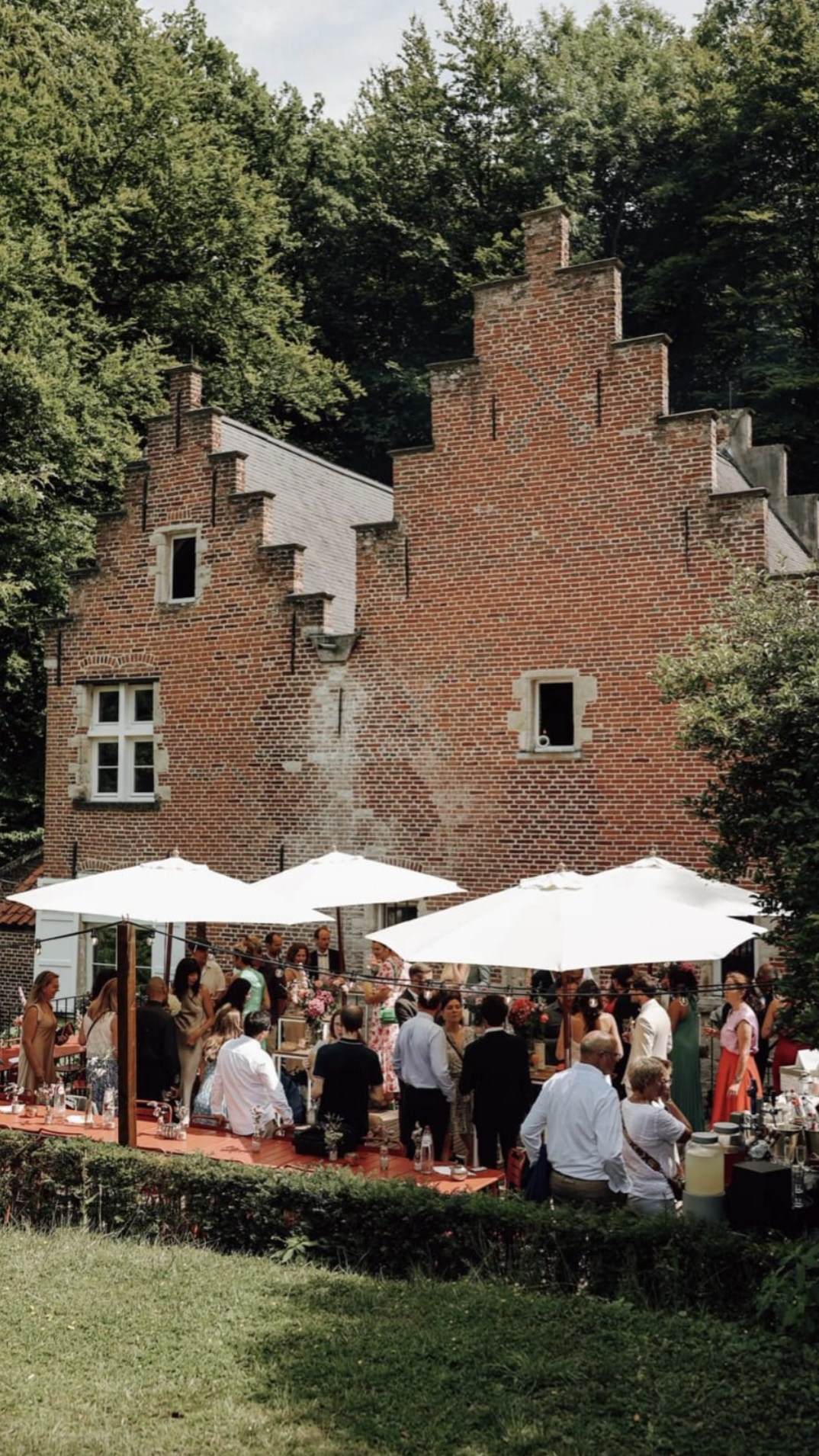 People gathered outdoors for an event under white umbrellas in front of the Spaans Huis surrounded by trees.