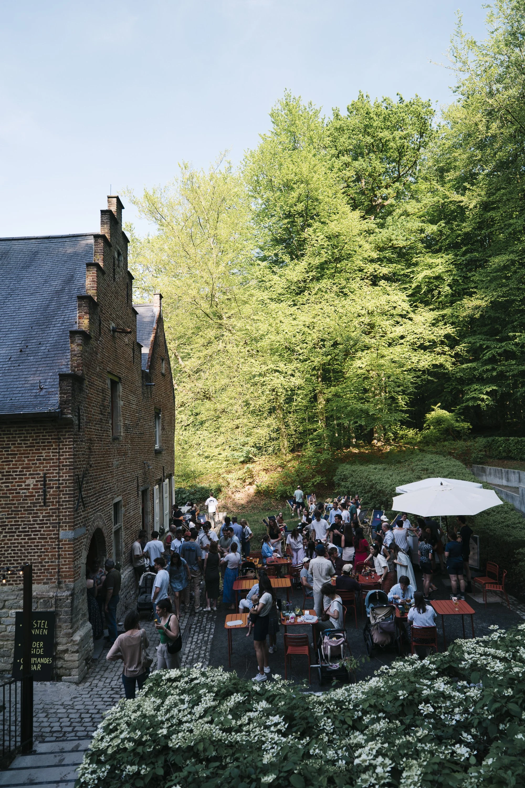 Outdoor gathering with many people near a brick building under large green trees, with tables, chairs, and a white umbrella.  At Spaans Huis - Tervuren