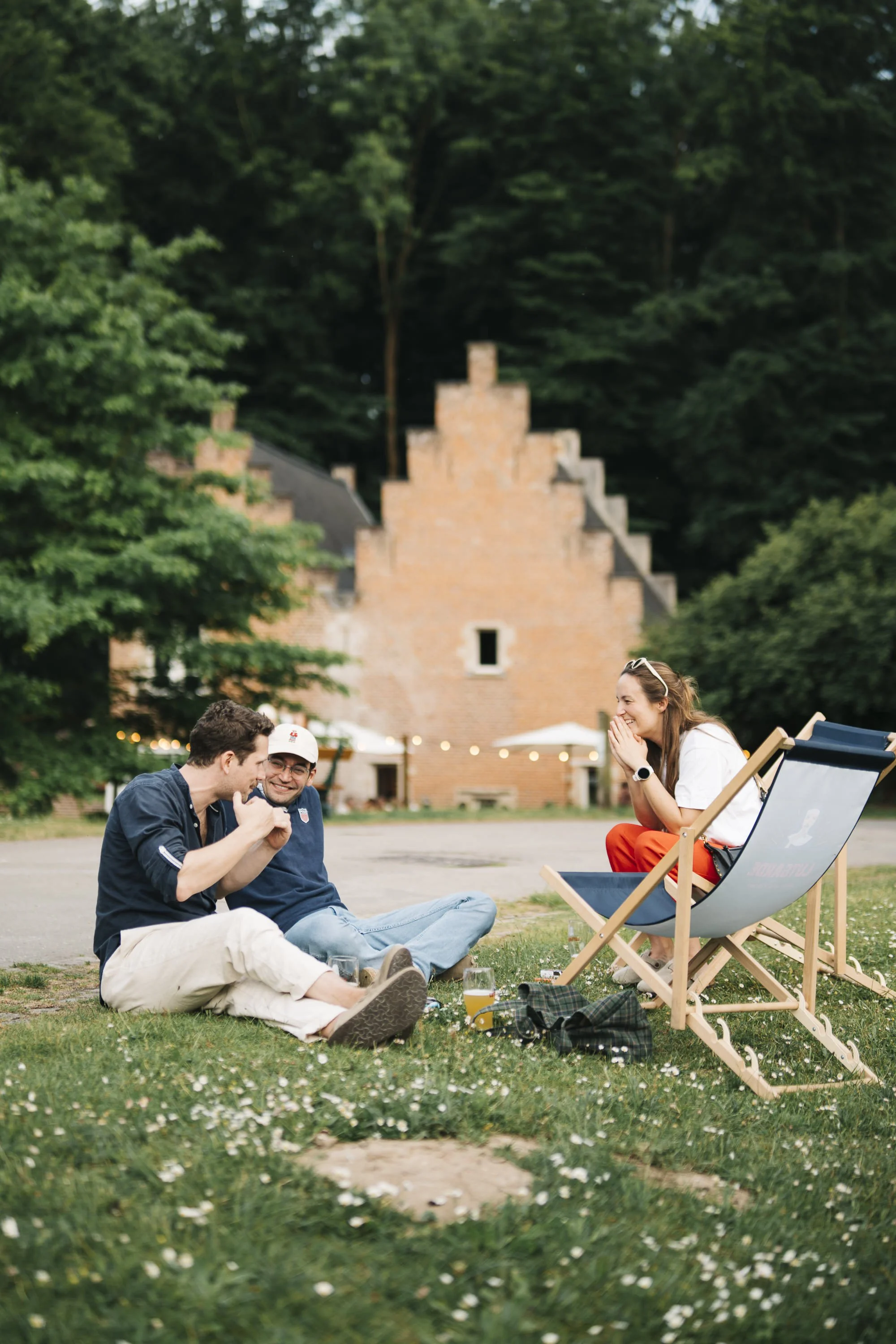 Three friends sitting and laughing on grass in front of a historic brick building (at Spaans Huis) with trees in the background. One friend is seated in a deck chair, the other two are sitting on the ground; they appear to be enjoying a picnic.