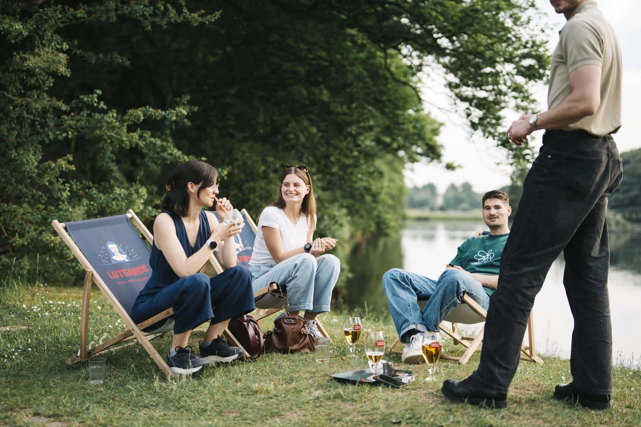 Group of four friends relaxing outdoors by the river, sitting on deck chairs, with drinks, in a lush green setting. At Spaans Huis Tervuren.