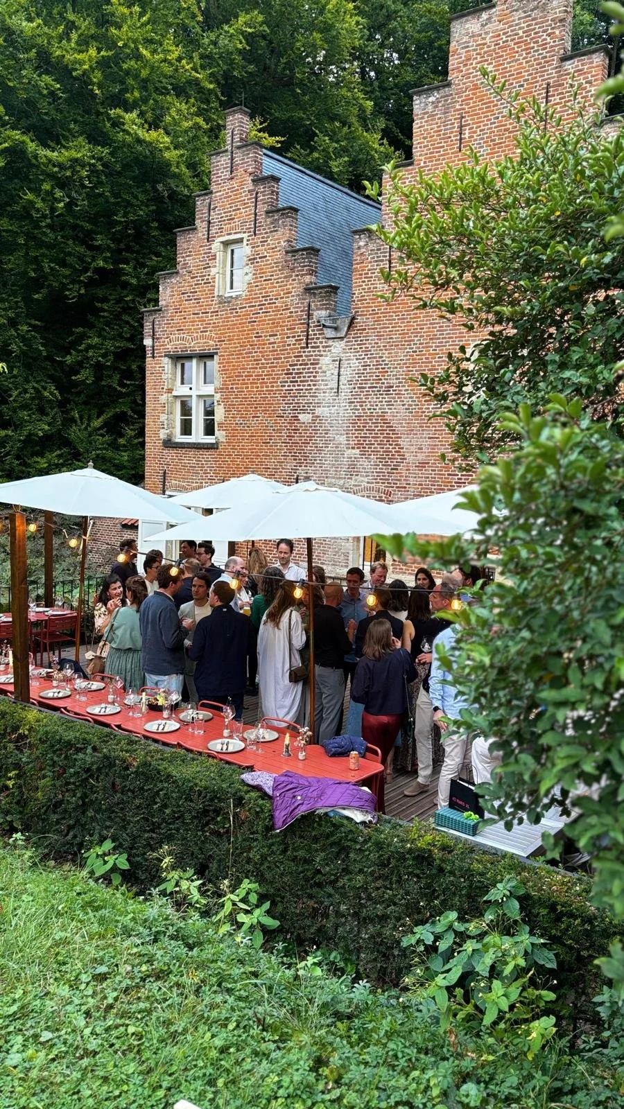 People gathered at an outdoor dinner party with tables, umbrellas, and string lights in front of an old brick building surrounded by greenery. At Spaans Huis Tervuren.