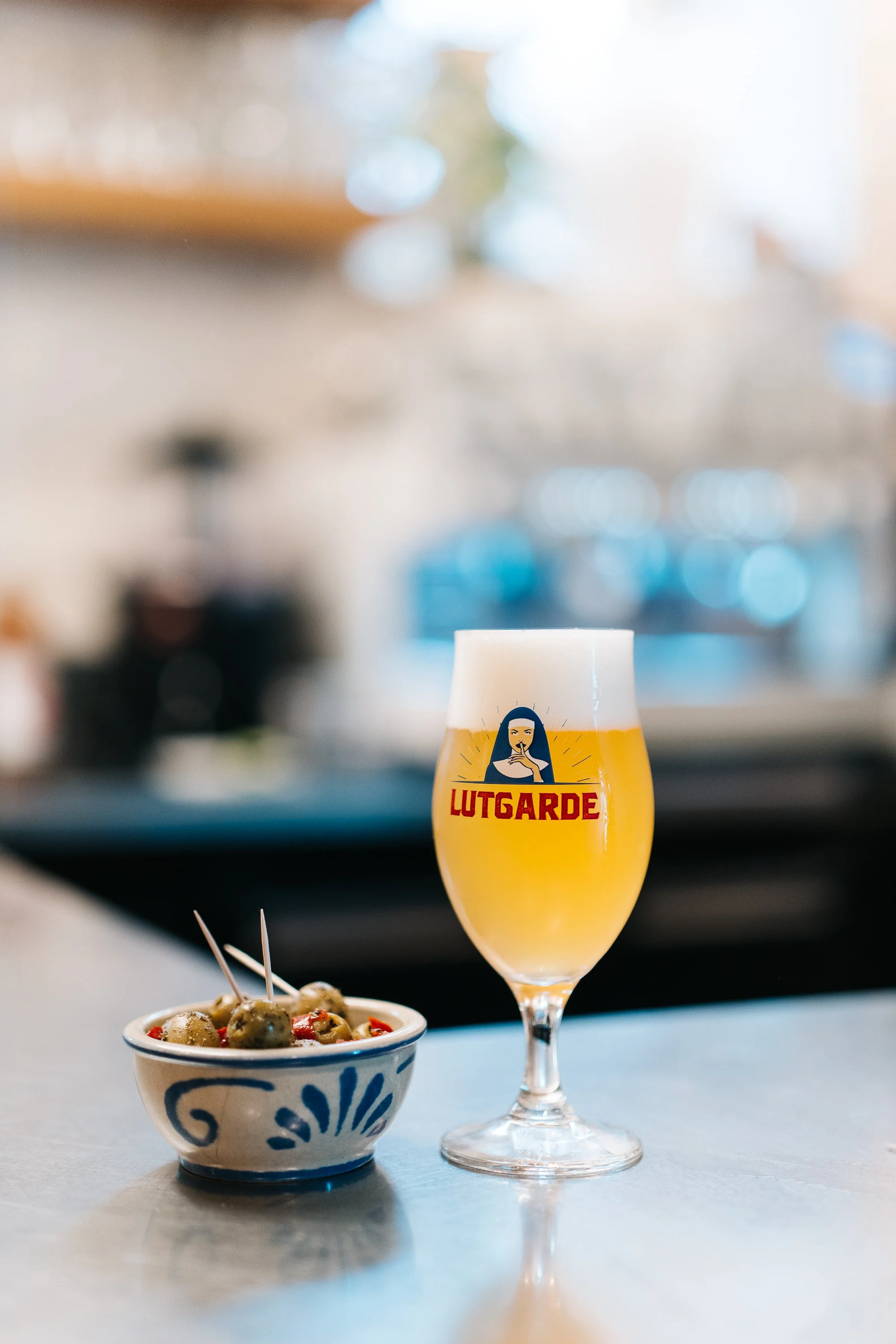 A glass of Witgarde beer with a foamy head next to a bowl of green stuffed olives with toothpicks on a light-colored table in a blurred indoor setting.