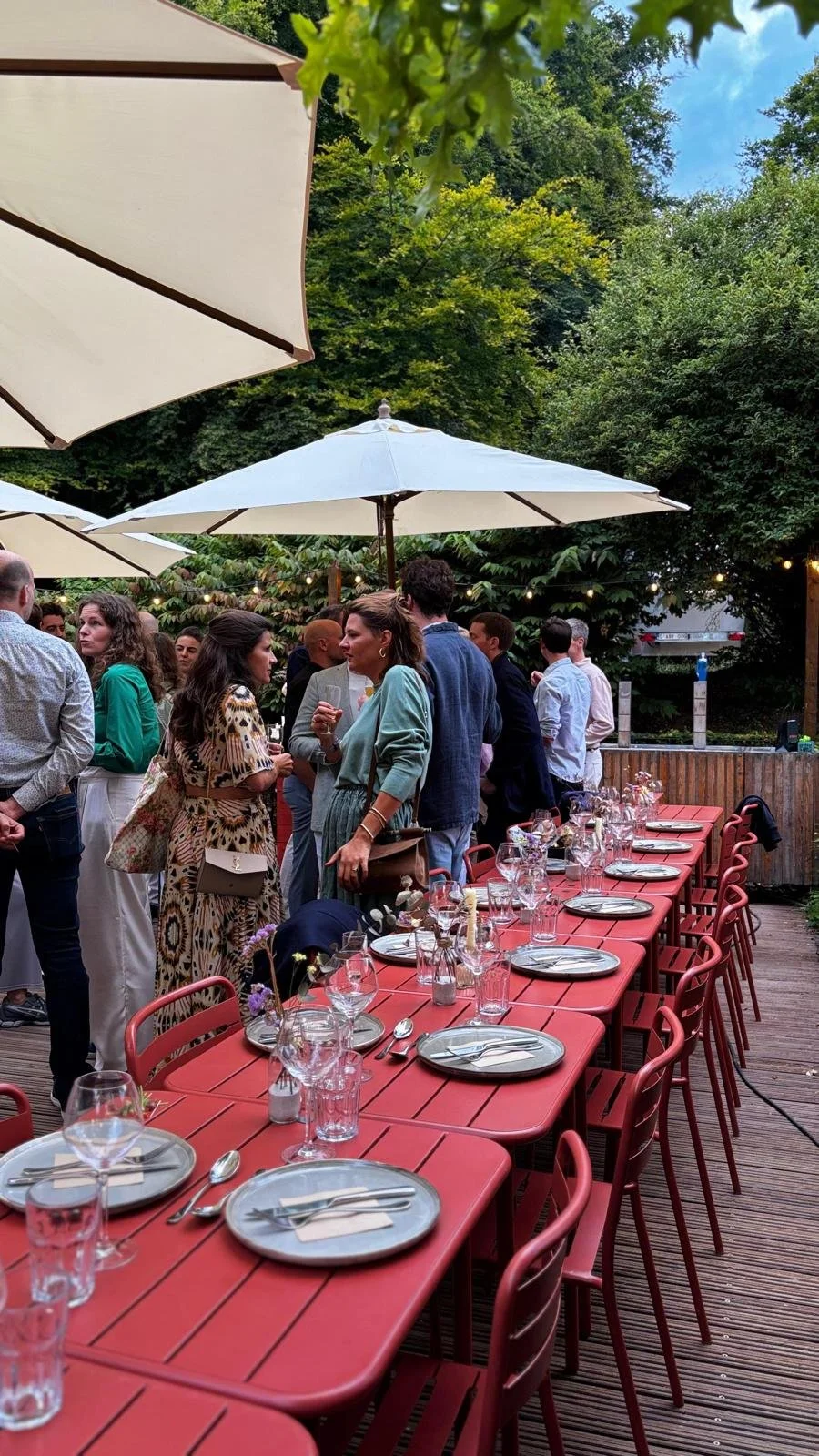 People socializing at an outdoor dining event with tables set with plates, glasses, and utensils, under large umbrellas surrounded by greenery. At Spaans Huis Tervuren.