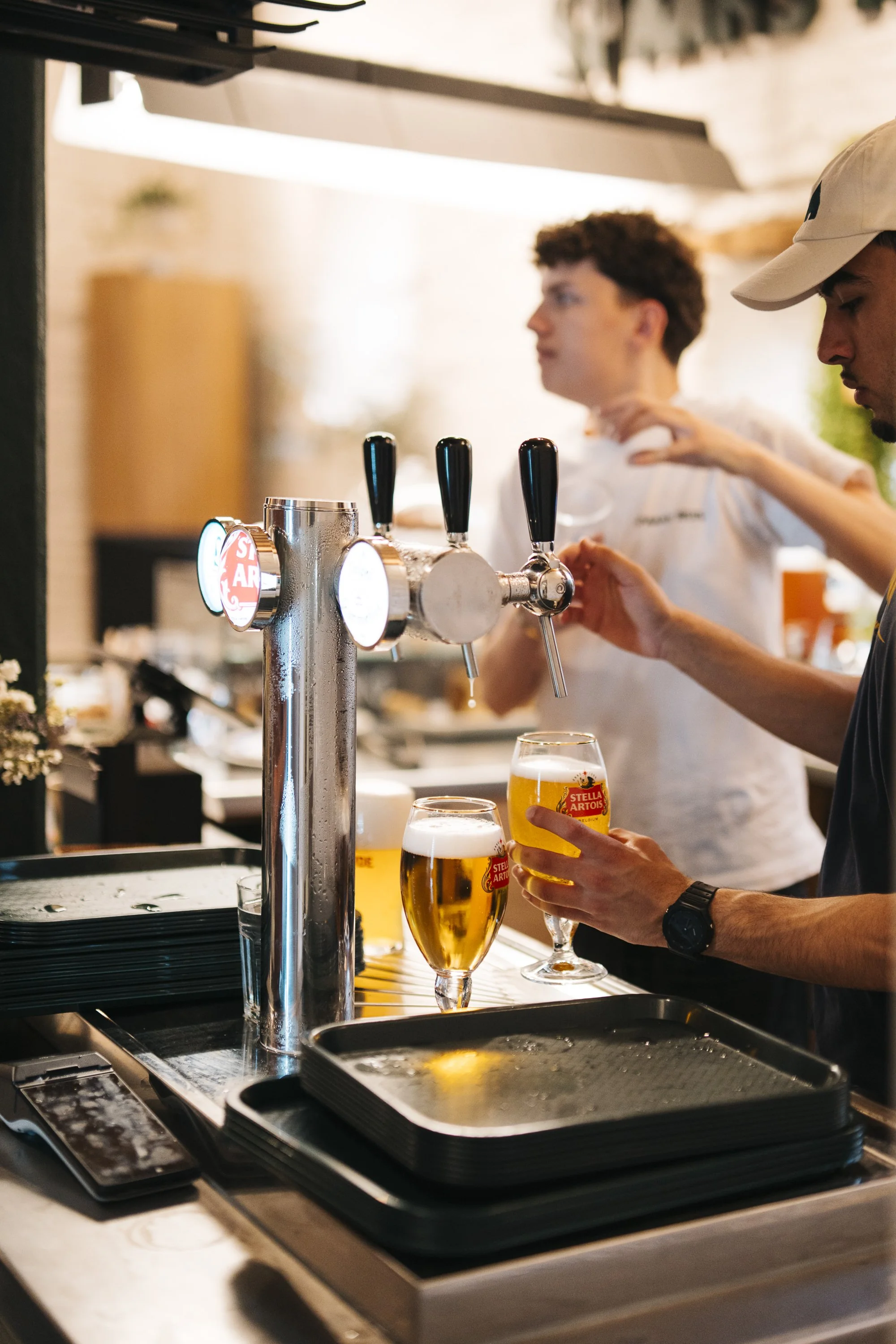 A bartender pours beer from a tap into a glass, with another glass already filled on the counter. The background shows a person in a white shirt with curly hair, and the counter has black trays, a smartphone, and a menu, indicating a bar or brewery s