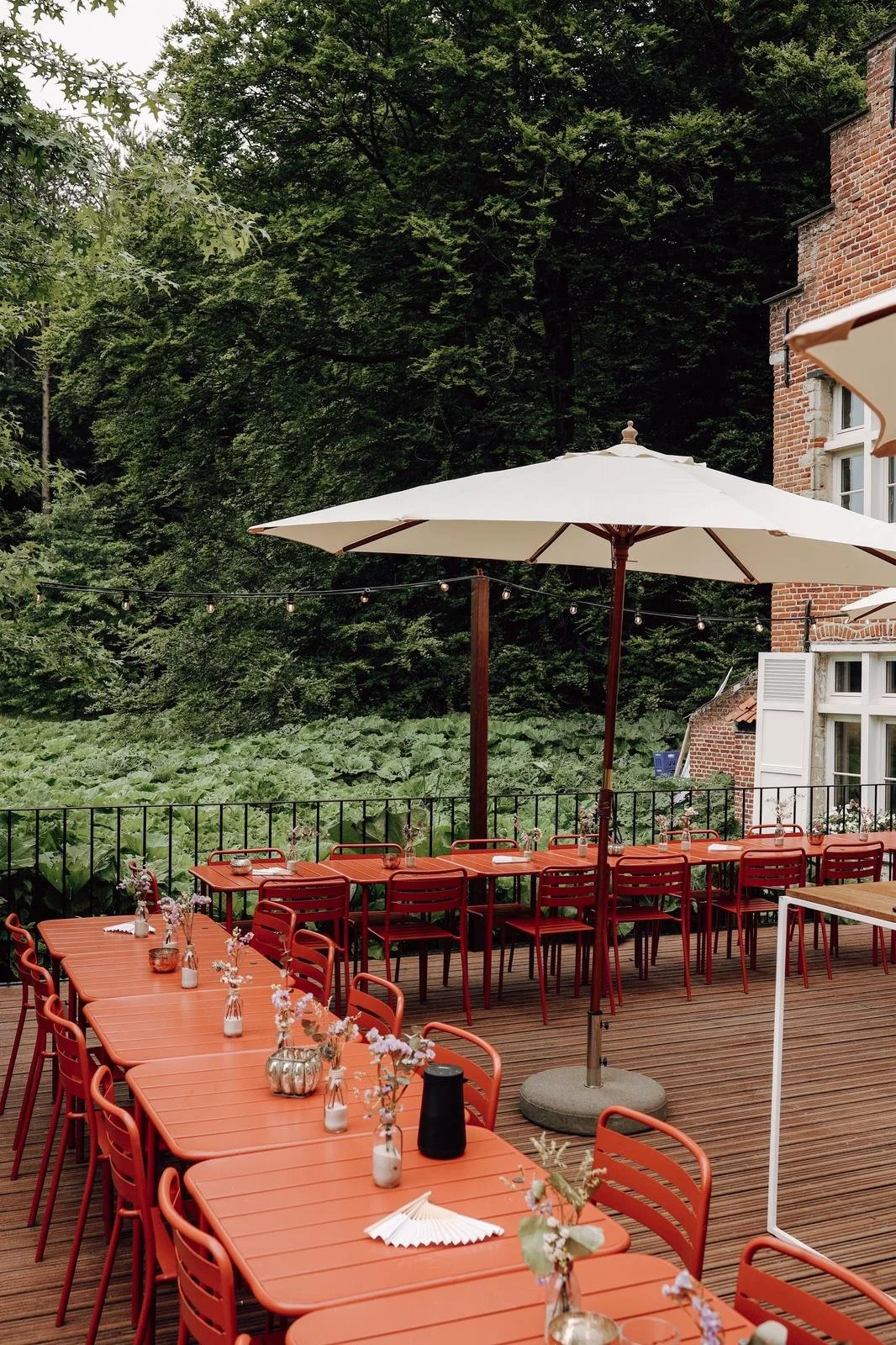 Outdoor dining area with red tables and chairs, umbrellas, and small vases with flowers, set on a wooden deck surrounded by greenery and trees.