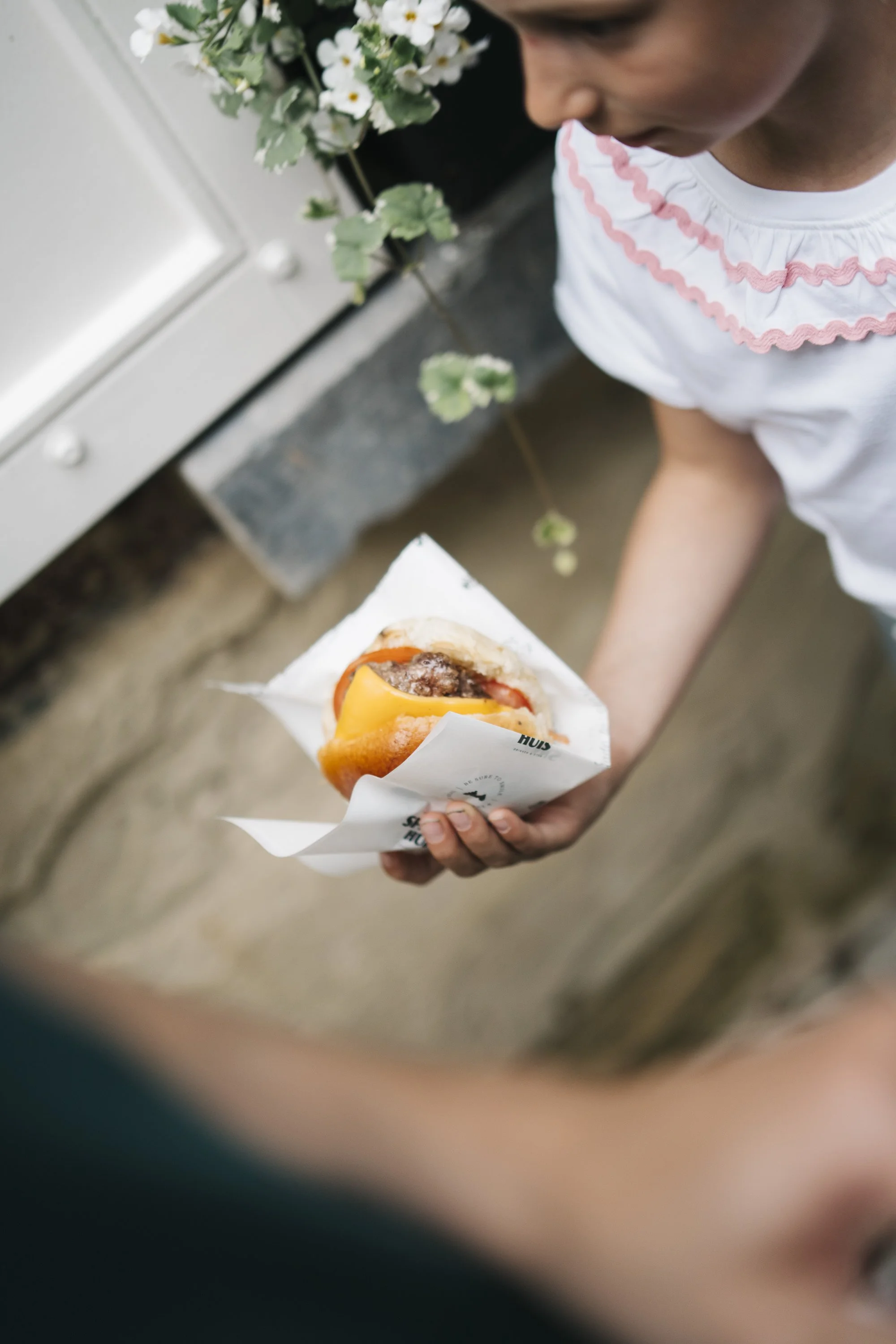 A young girl holding a wrapped hamburger with cheese and toppings, outdoors near a white door and potted plant.  At Spaans Huis - Tervuren.