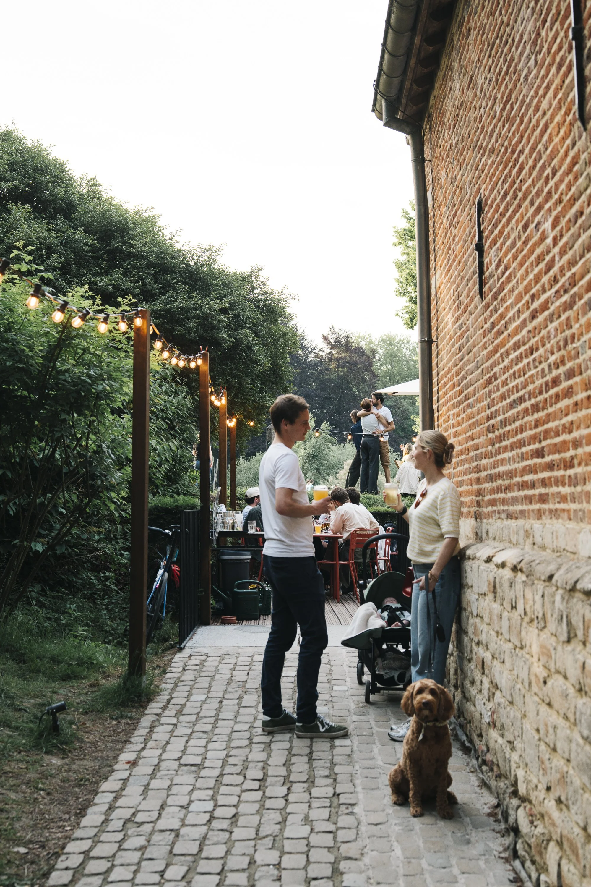 People socializing outside on a cobblestone pathway at sunset, with string lights overhead. A woman with a dog and a baby stroller is talking to a man holding a drink, near a brick building.  at Spaans Huis - Tervuren.