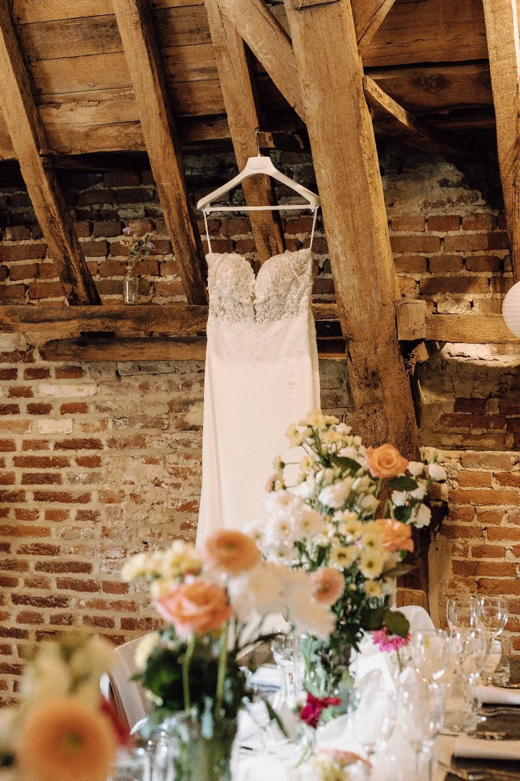 A wedding dress hanging on a wooden beam in a rustic setting with brick walls and floral arrangements on a dining table in the foreground. At Spaans Huis Tervuren.