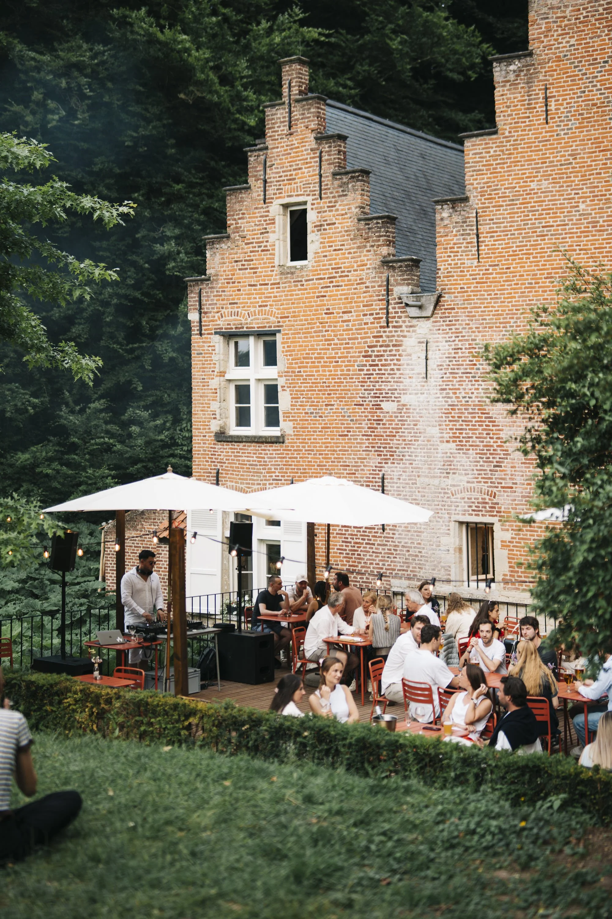 Outdoor gathering with people dining at tables on a patio beside a brick building, surrounded by greenery and trees, during the evening. At Spaans Huis Tervuren.
