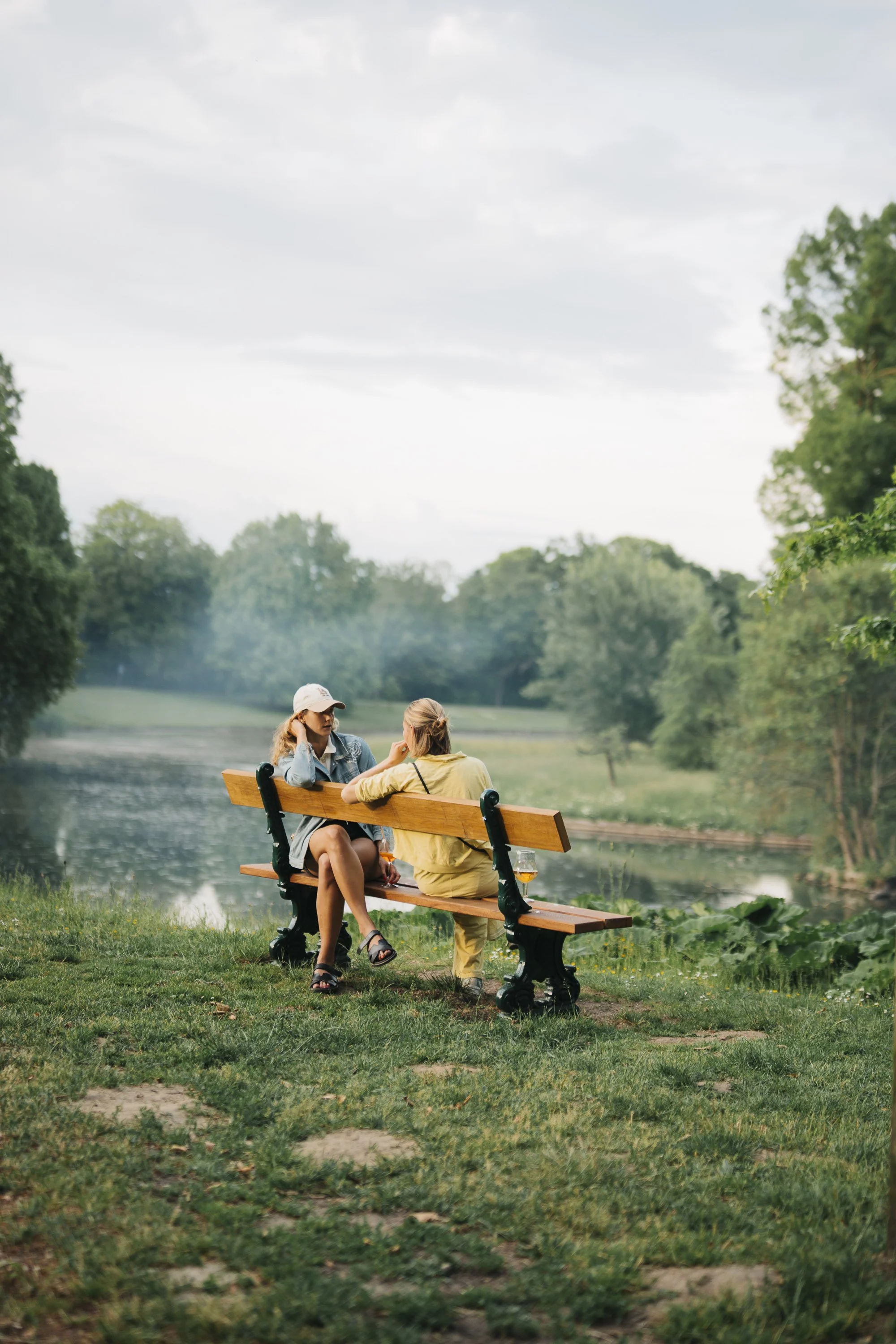 Two women sitting on a wooden park bench near a pond, engaged in conversation, surrounded by trees and greenery near the Spaans Huis.