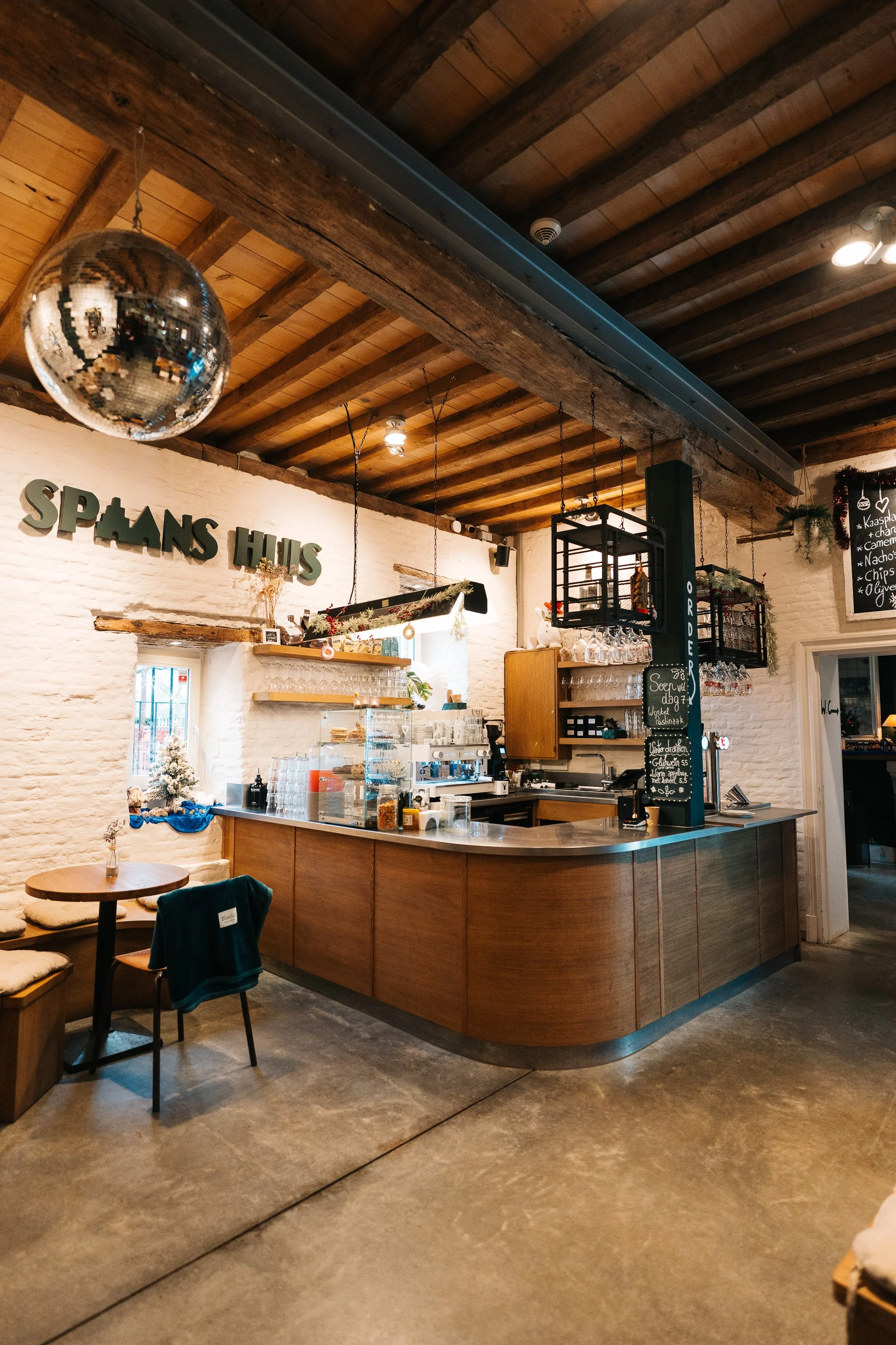 Cozy cafe with a curved wooden counter, white brick walls, and warm wood ceiling beams. Small table with chairs, hanging glassware, and a disco ball hanging from the ceiling, with "SPAANS HUS" sign on the wall.