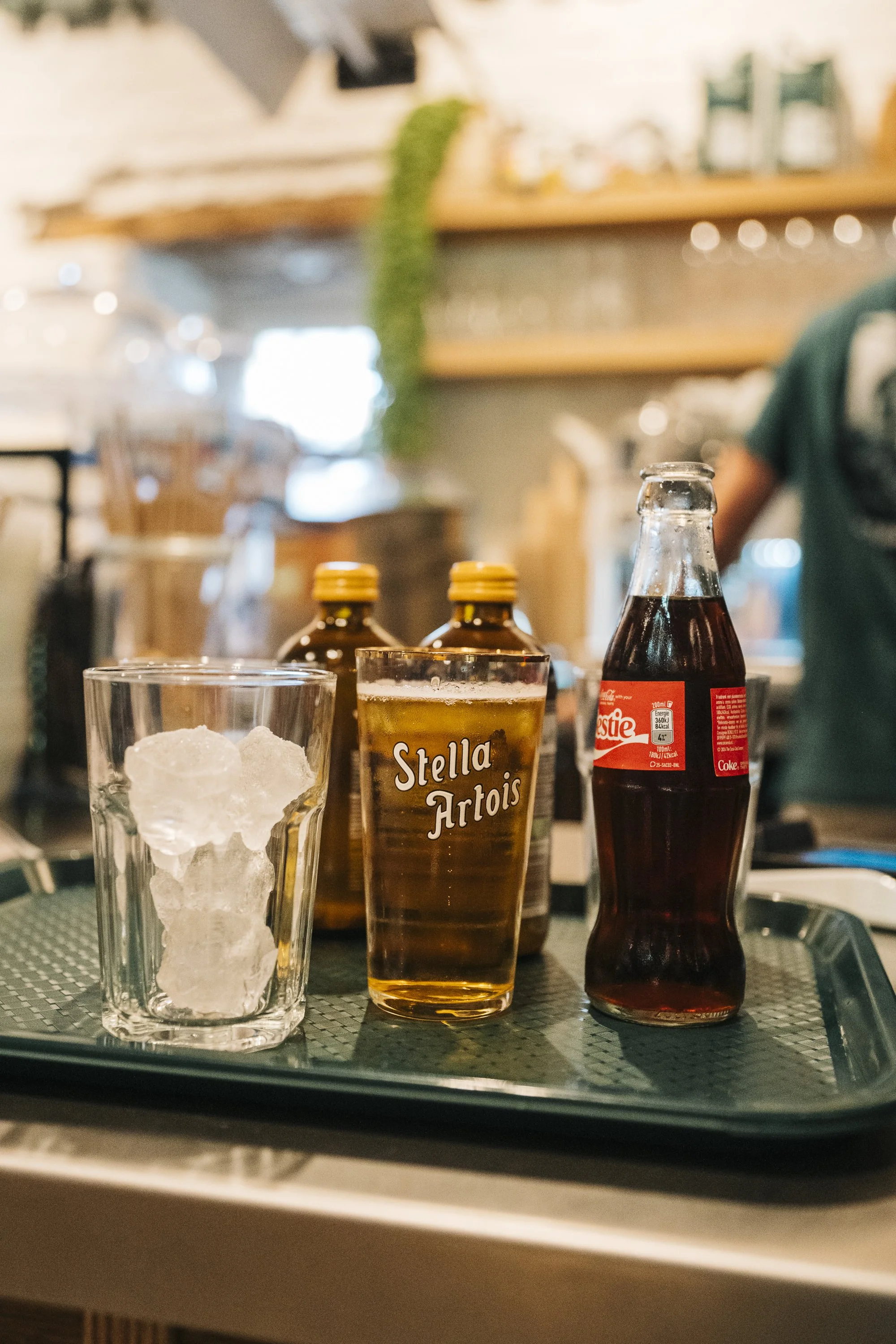 A tray with a glass of ice, a glass of Stella Artois beer, a bottle of cola, and two bottles of pills on a counter at Spaans Huis - Tervuren