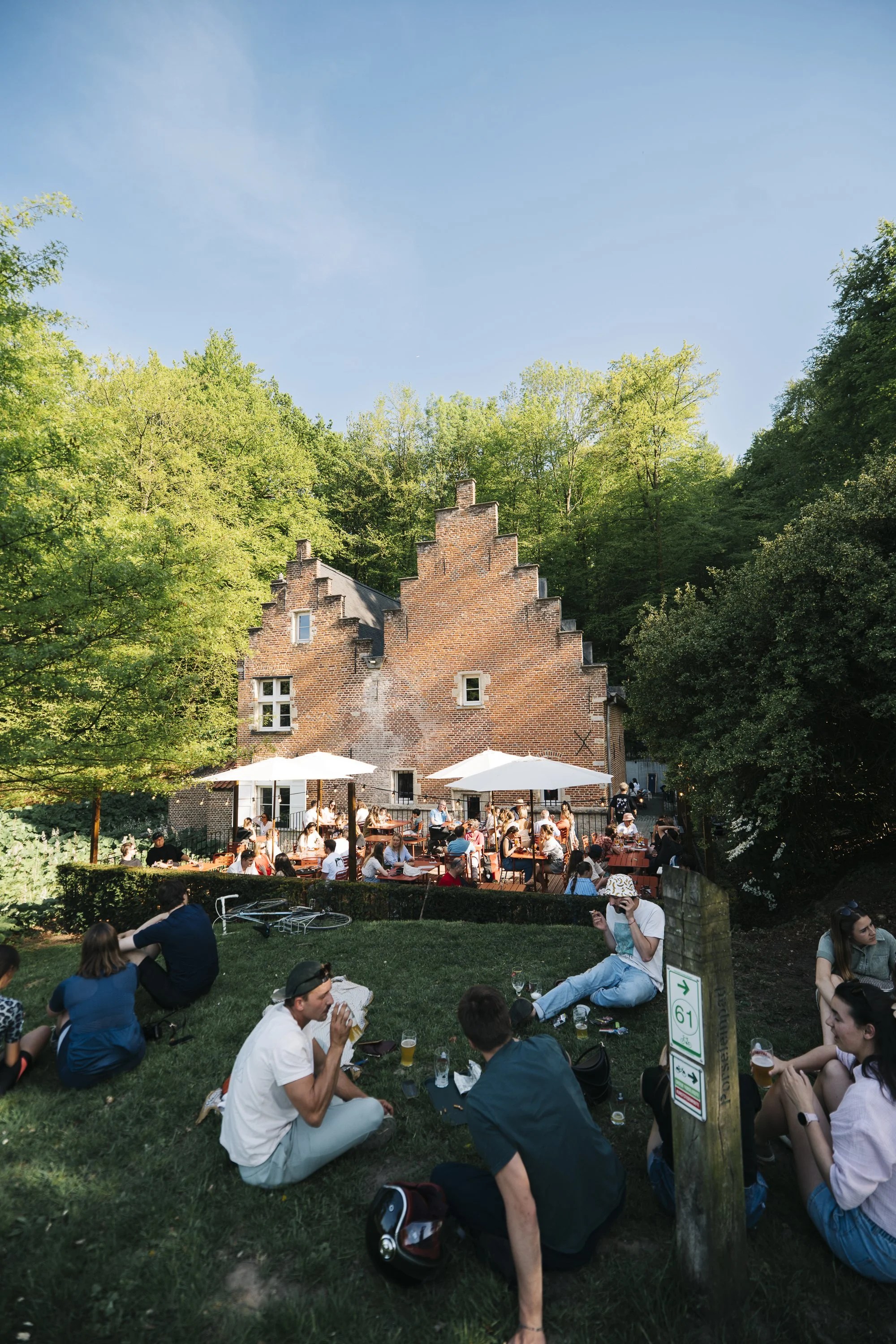 People enjoying a picnic and outdoor dining on grass and under umbrellas in front of a brick house surrounded by trees on a sunny day.  at Spaans Huis - Tervuren.