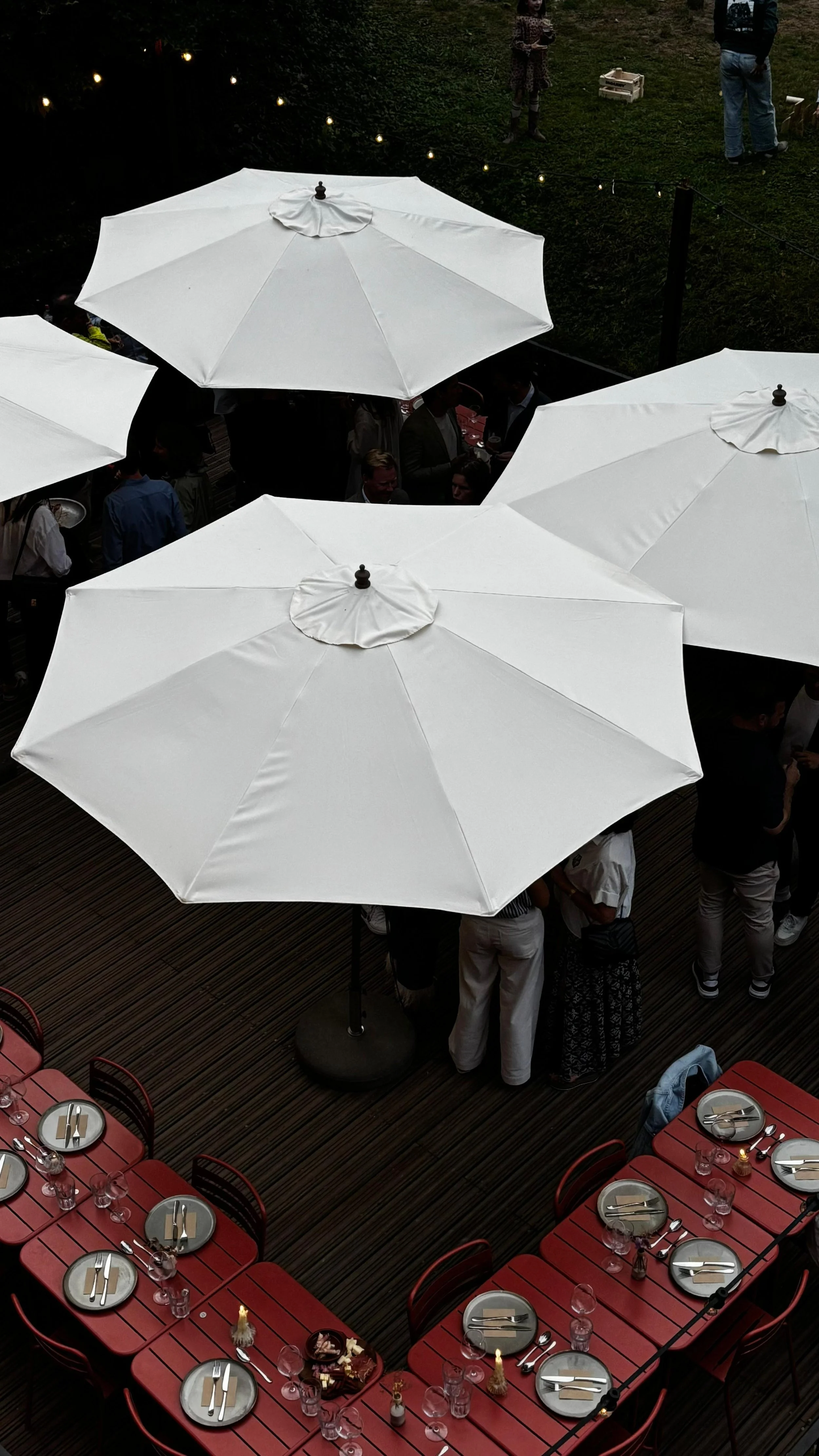 Top view of an outdoor dining area with red tables, gray plates, silverware, glasses, and candles, surrounded by large white patio umbrellas and people socializing underneath. At Spaans Huis Tervuren.