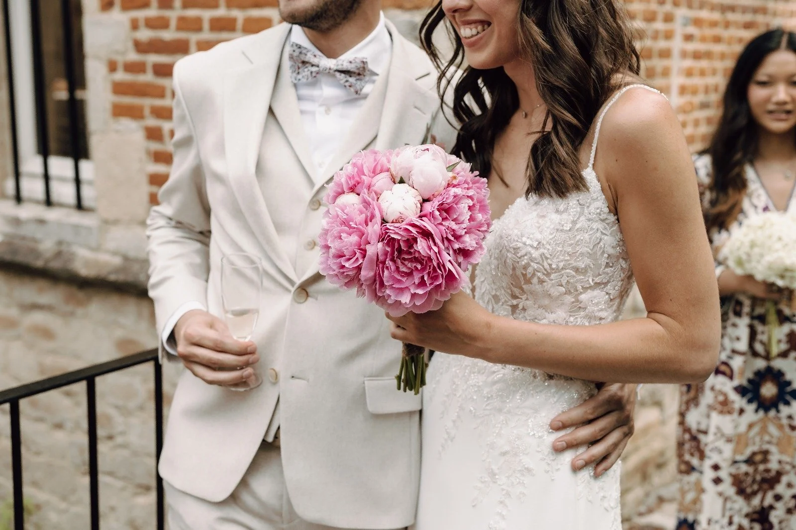 Bride holding a pink floral bouquet at a wedding ceremony, wearing a lace wedding dress, standing close to the groom dressed in a light-colored suit, with a woman in the background holding a bouquet. At Spaans Huis Tervuren.