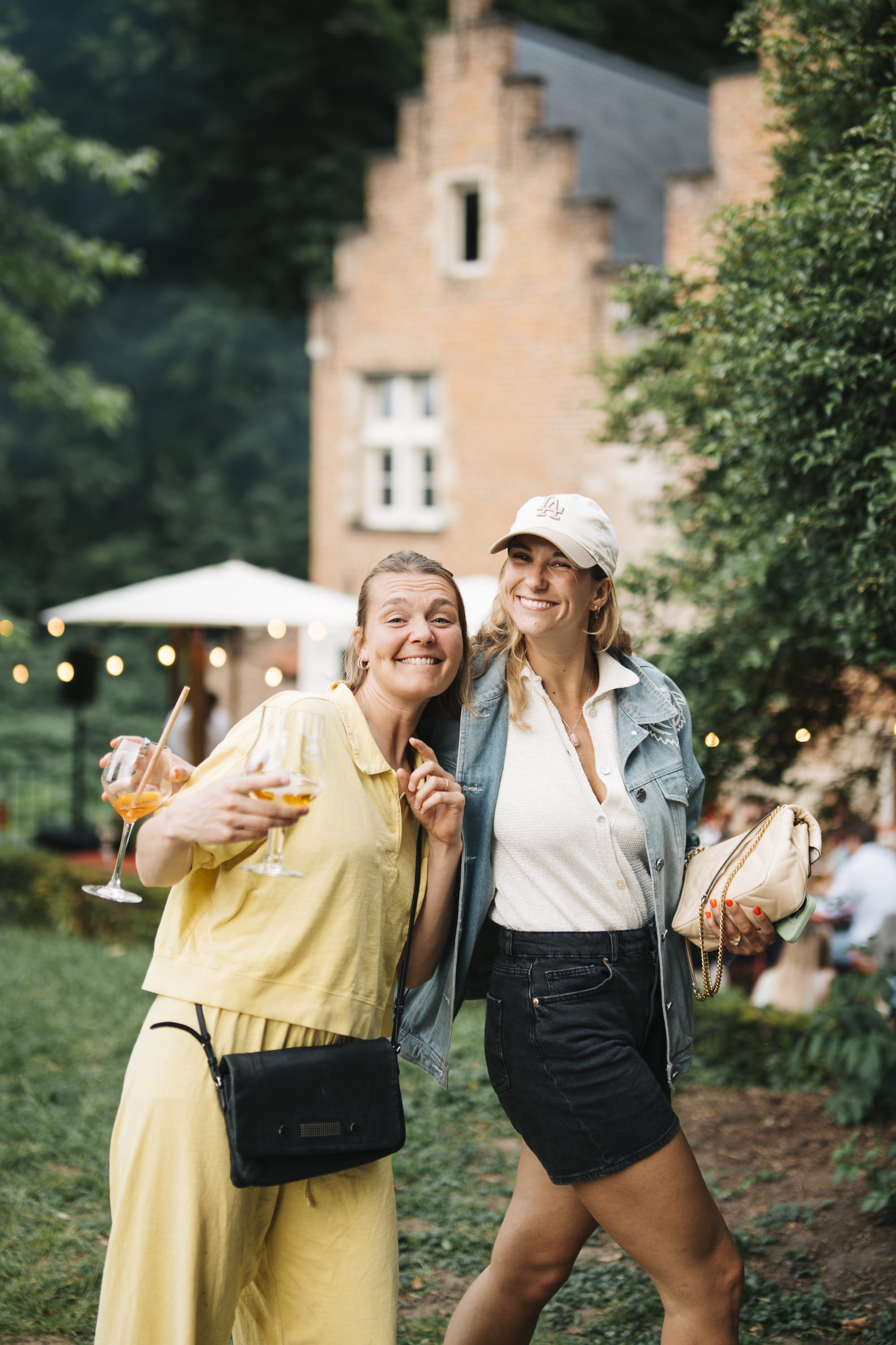 Two women smiling and posing at an outdoor gathering, holding drinks, with a large house and trees in the background. At Spaans Huis Tervuren.