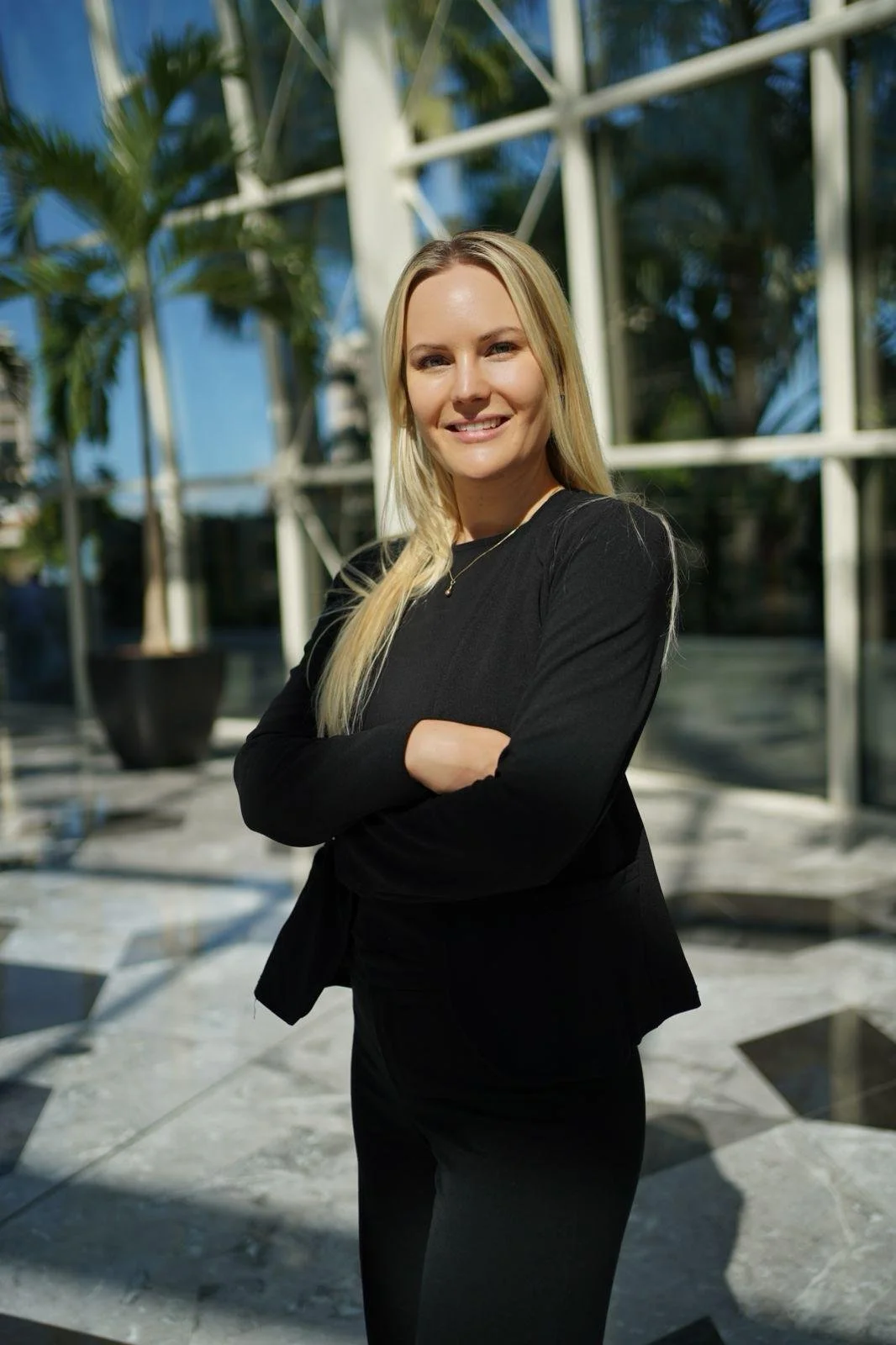 A blonde woman in a black outfit with arms crossed, smiling outdoors with a modern geometric glass building and palm trees in the background.