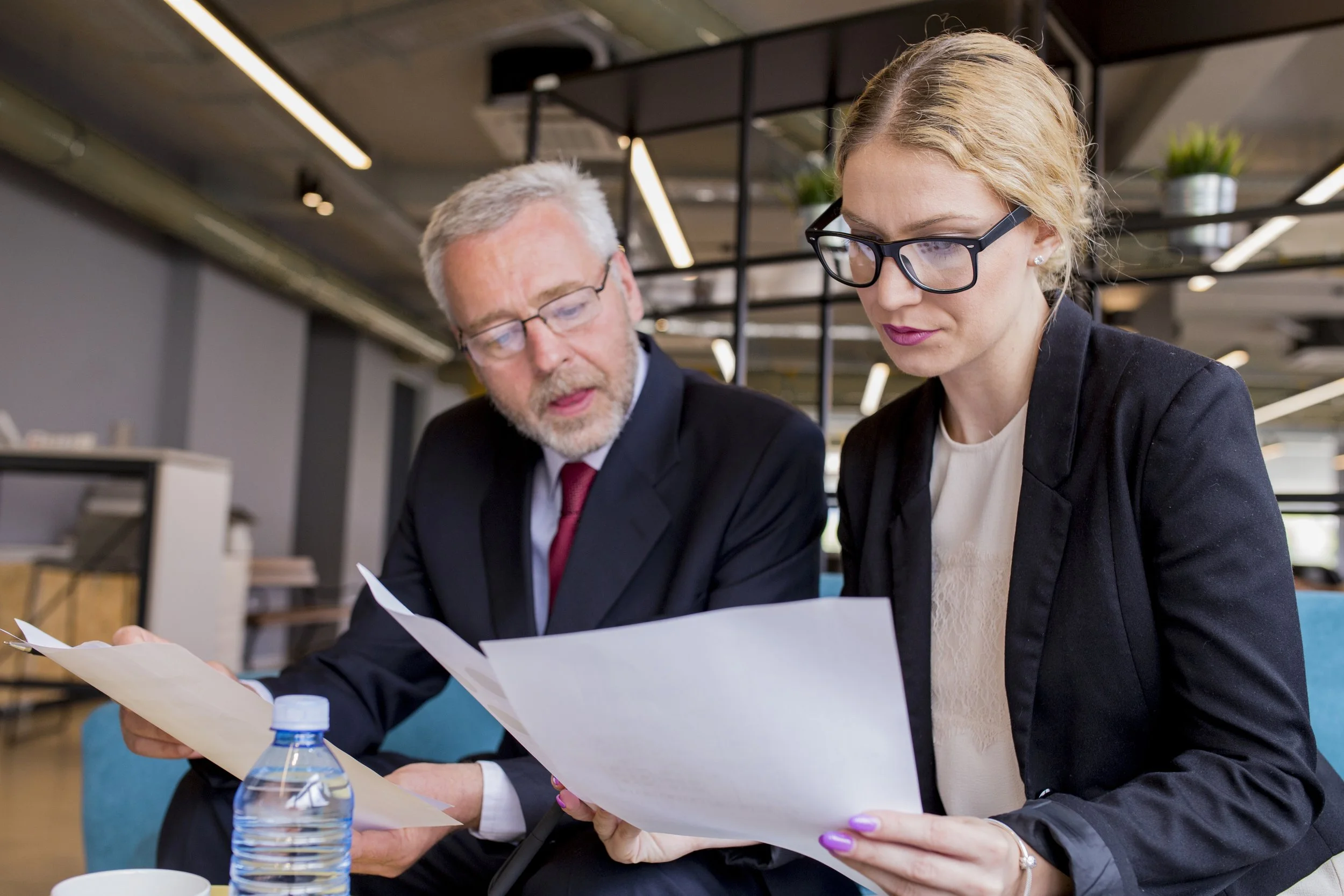 Two professionals, a man and a woman, sitting at a table in an office or conference area, looking at documents together.