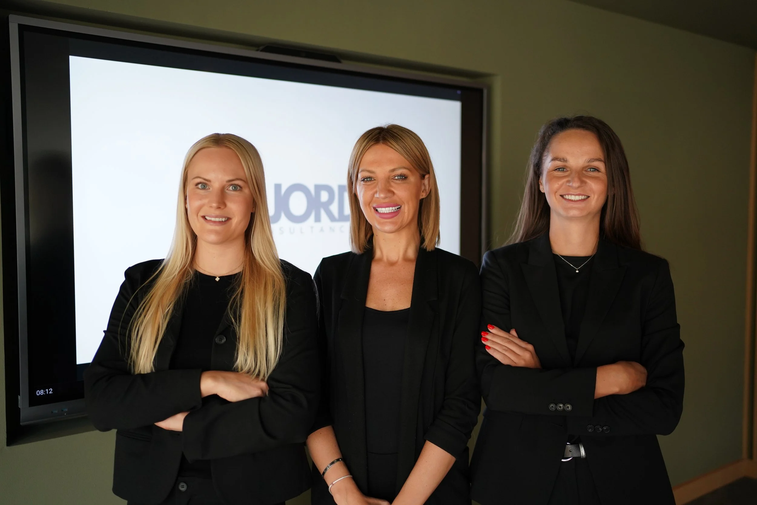 Three women standing together indoors in front of a screen, smiling, dressed in black professional attire.