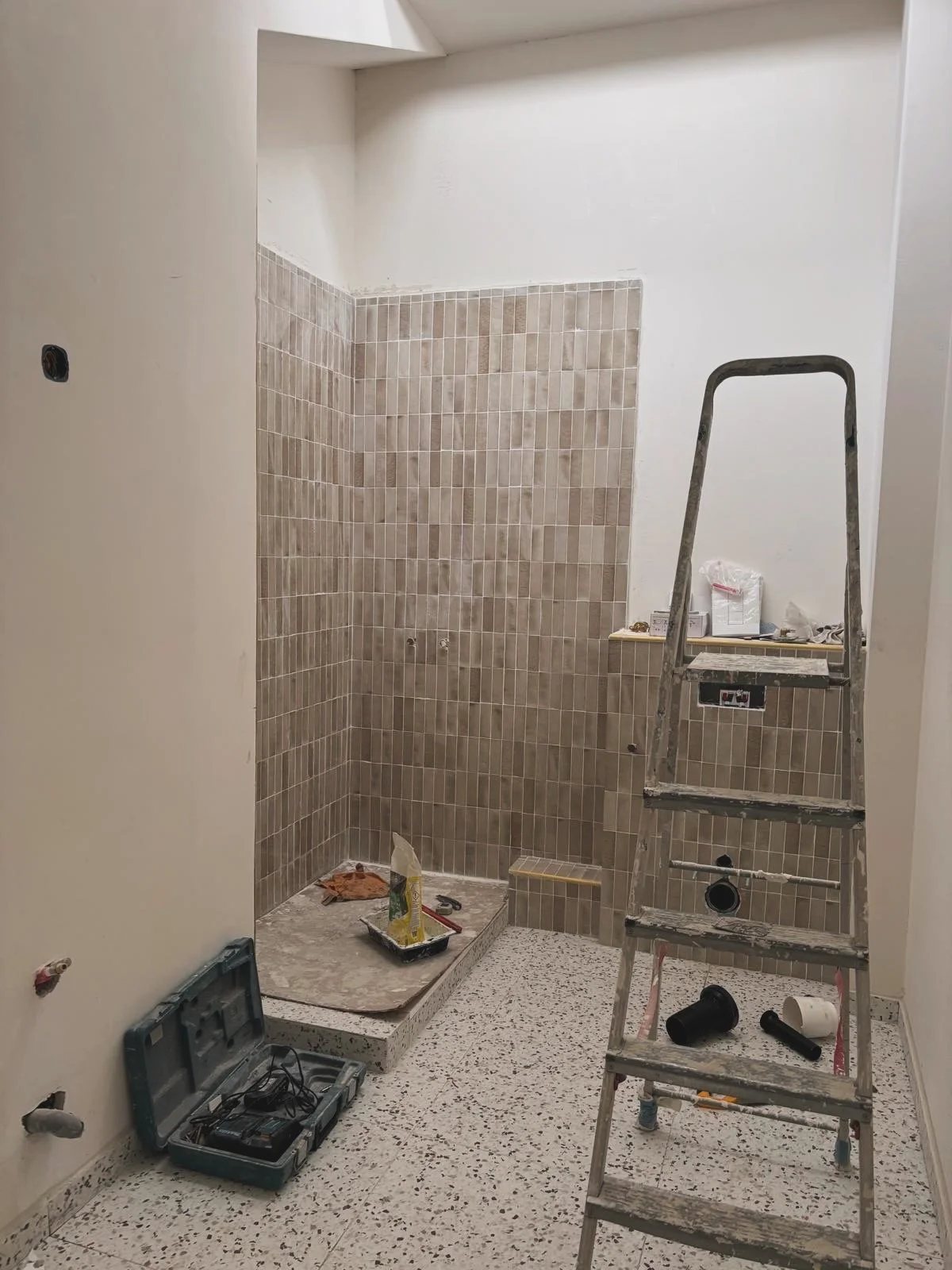 Une pièce en cours de rénovation avec un mur en carreaux beige et un sol en mosaïque. Il y a un escabeau en aluminium, des outils, un coffret à outils, et des matériaux de construction.