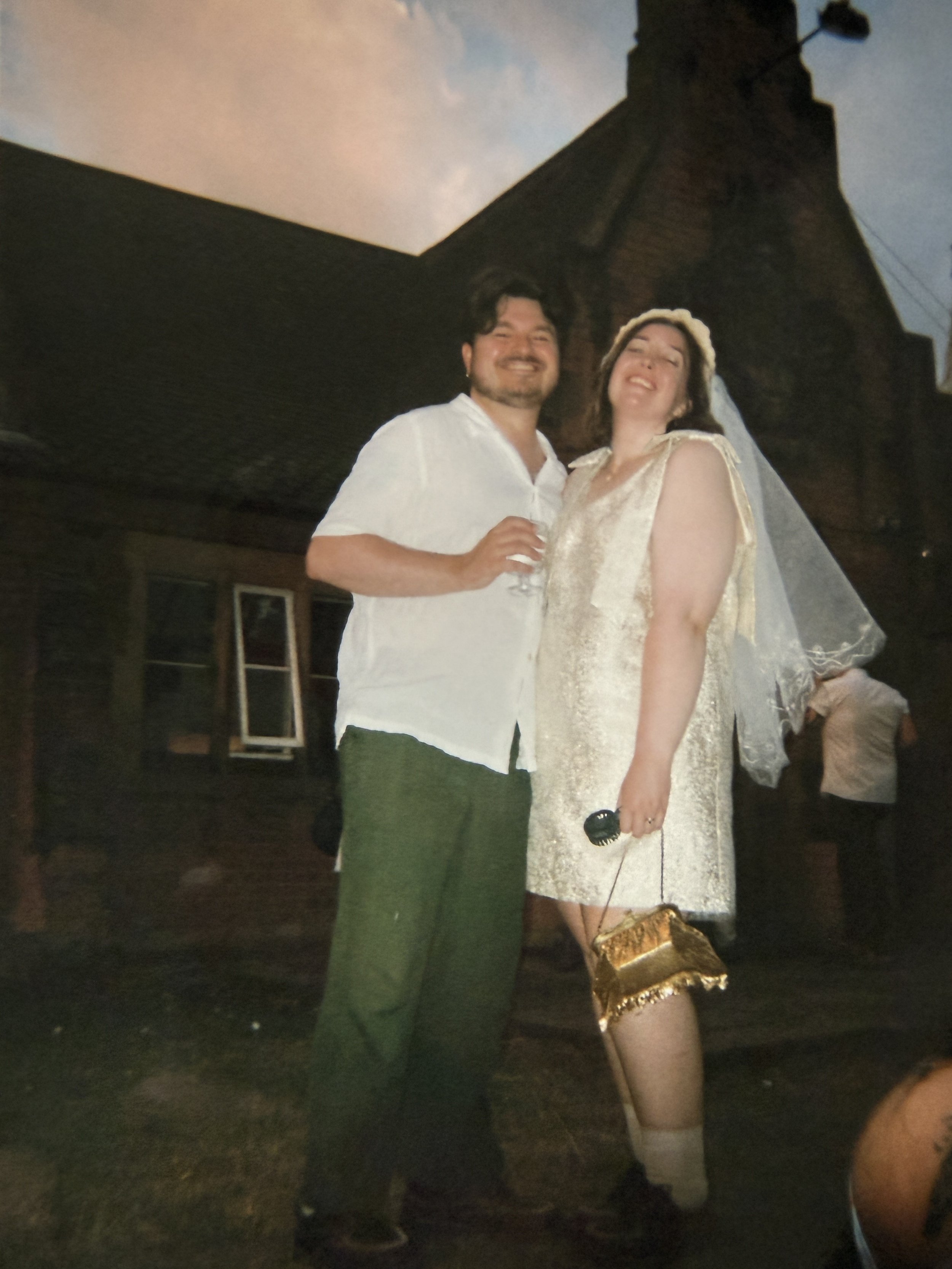 A bride and groom both smiling and posing for a photo. The man is holding a glass, and the woman is holding a small gold purse with a gold gown and veil. The background shows a dark outdoor setting with a building and a cloudy sky.