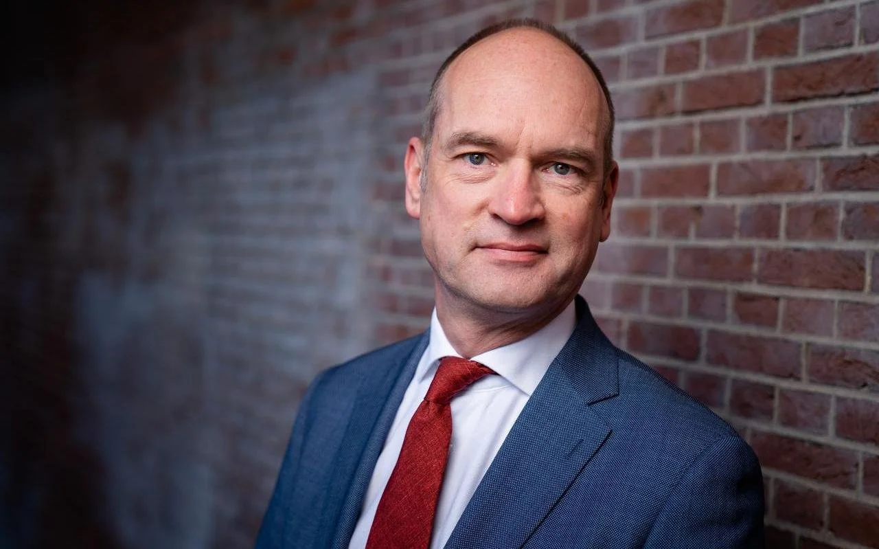 Portret van een man met bril en grijs haar, gekleed in een donker pak en witte overhemd, kijkend naar rechts
