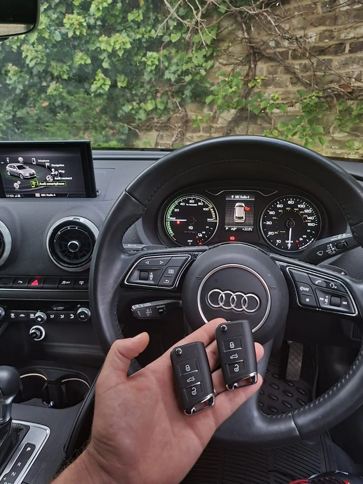 Ferson hånd holder to bilnøgler inde i en Audi, bagrunden viser bilens rat, instrumentbræt og et indbygget display i bilen.