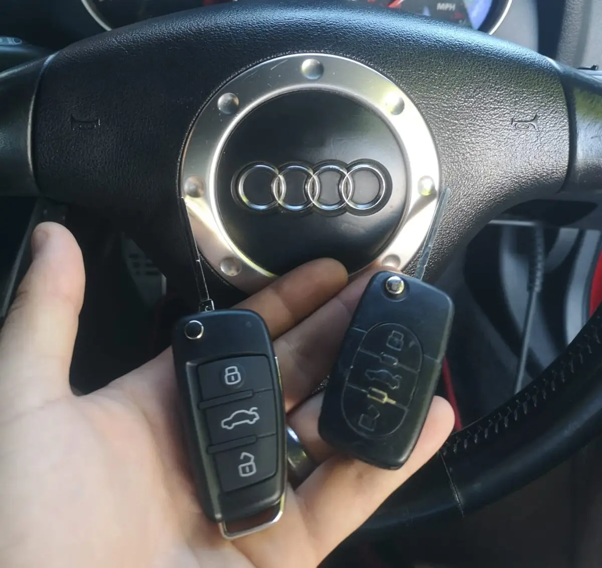 To bilnøgler holdes foran et Audi rat, som har det karakteristiske Audi-logo i midten.