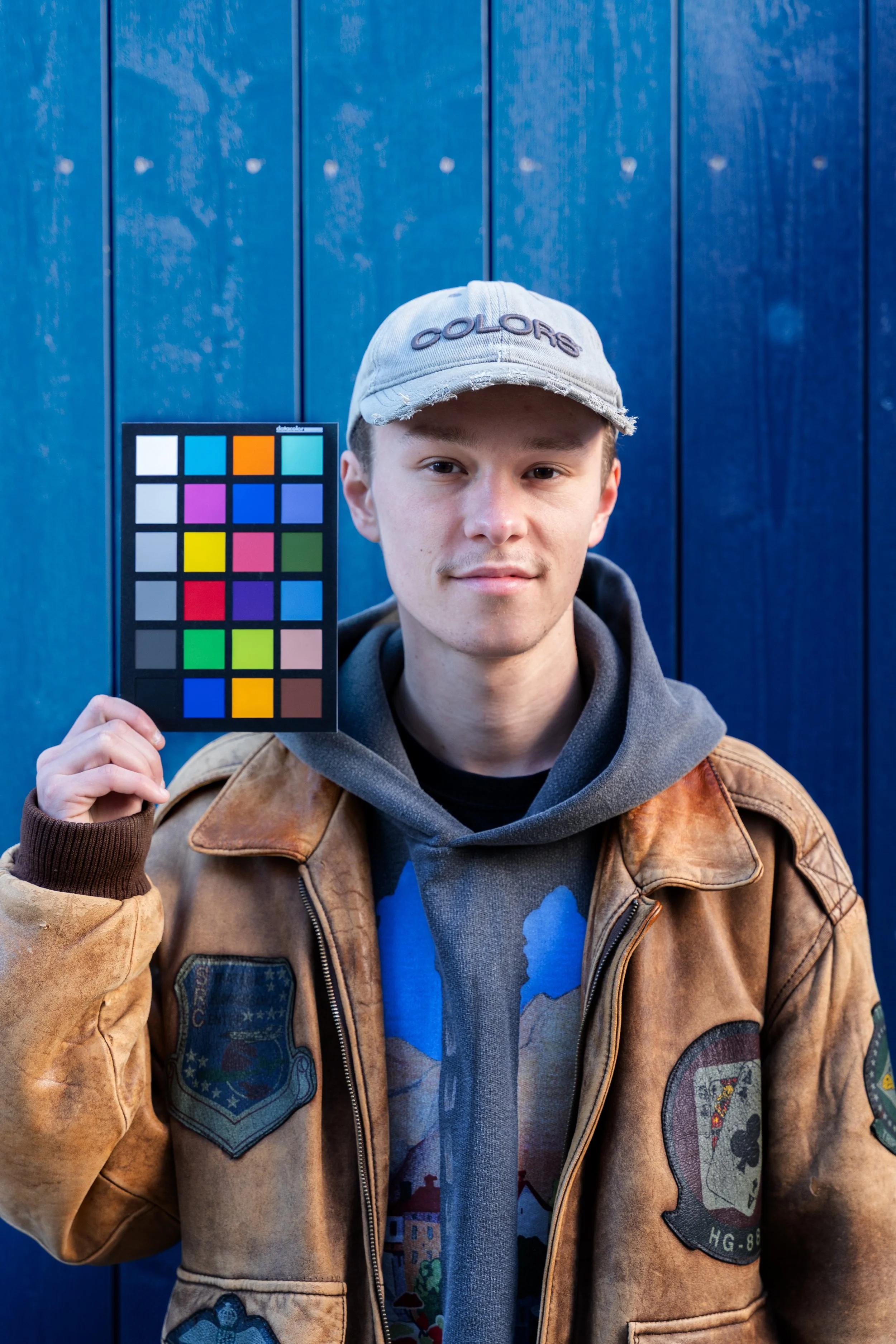 Junge Mann trägt grauen Kappe mit Aufschrift 'Colors' und braune Jacke mit Abzeichen, hält Farbvergleichskarte gegen blauen Holzhintergrund.