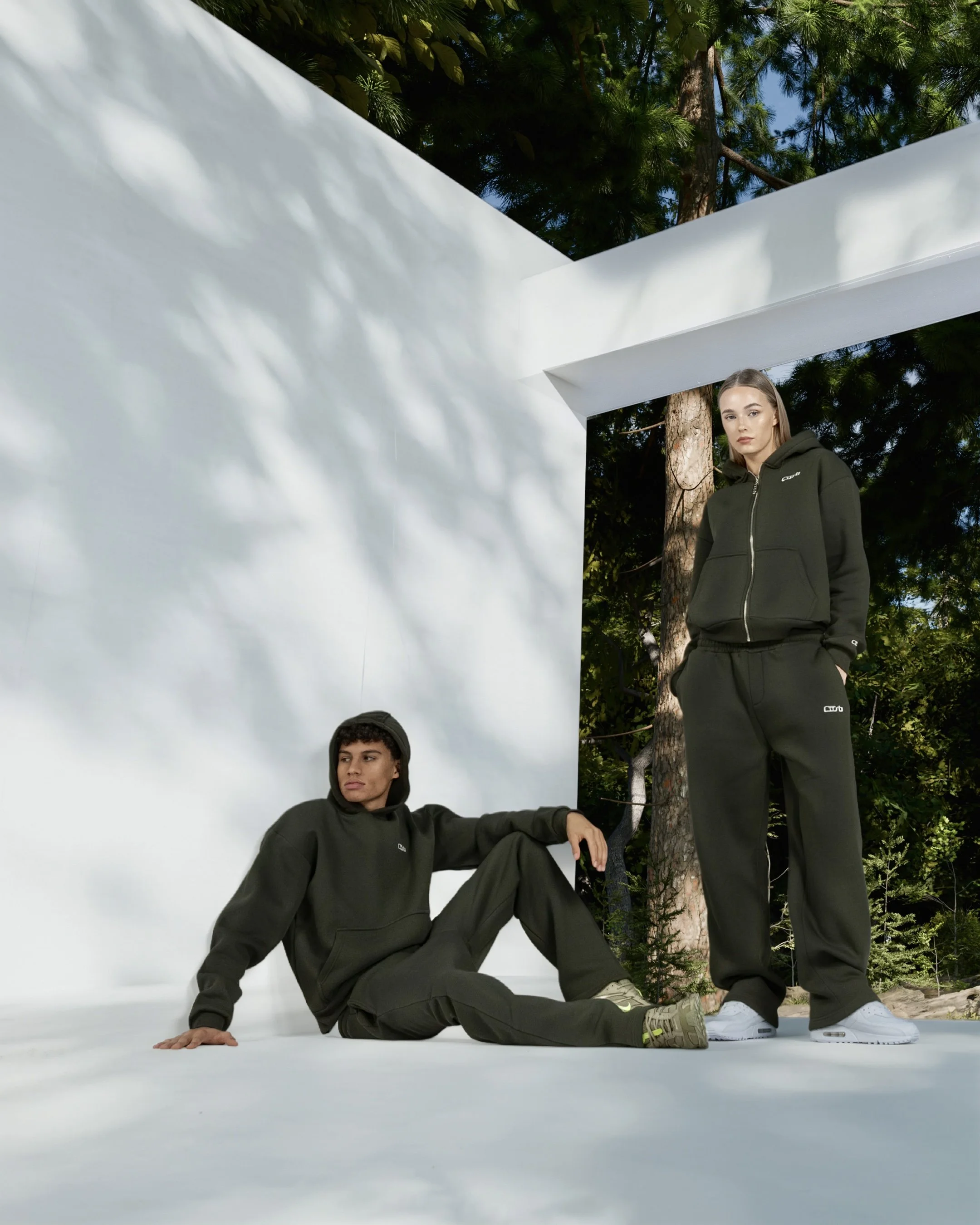 Zwei junge Erwachsene in CUSB Sport-Hoodies und Jogginghosen in einer Outdoor-Szene mit weißen Strukturen und Bäumen.