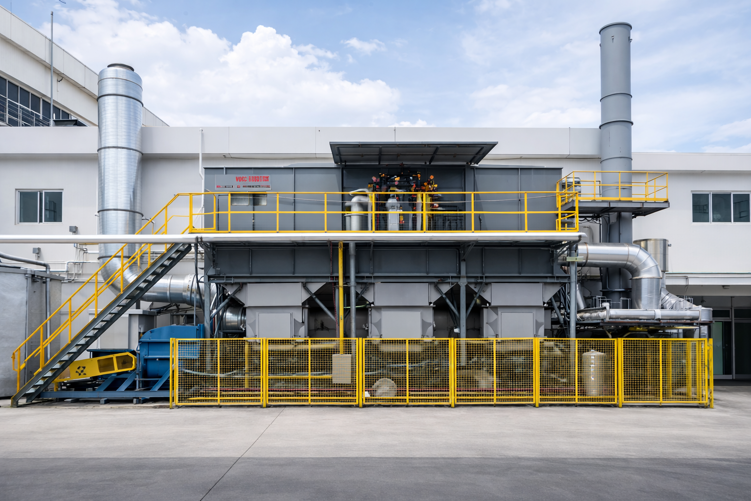Industrial equipment with yellow safety railings, stairs, pipes, and machinery outside a building under a partly cloudy sky.
