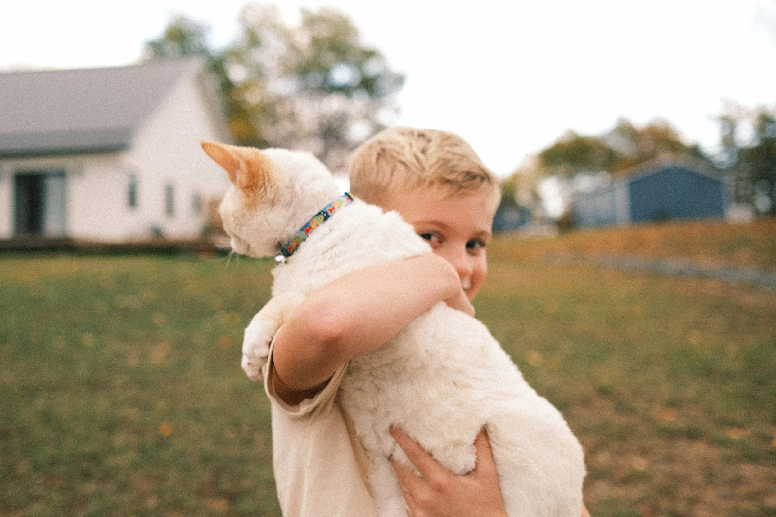 A young boy holding a white dog outdoors with a house and trees in the background.
