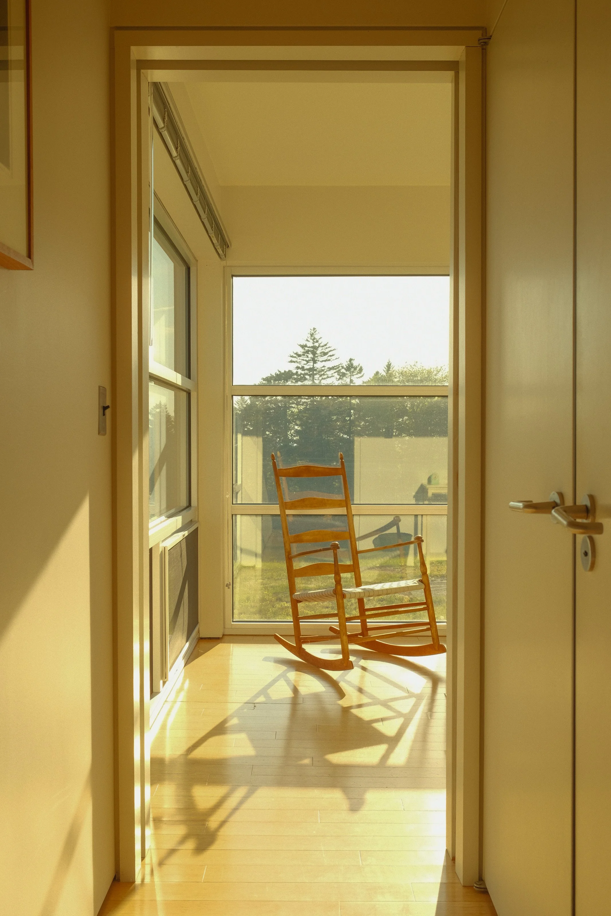 Sunlit room with a wooden rocking chair near large windows showing an outdoor landscape of trees.