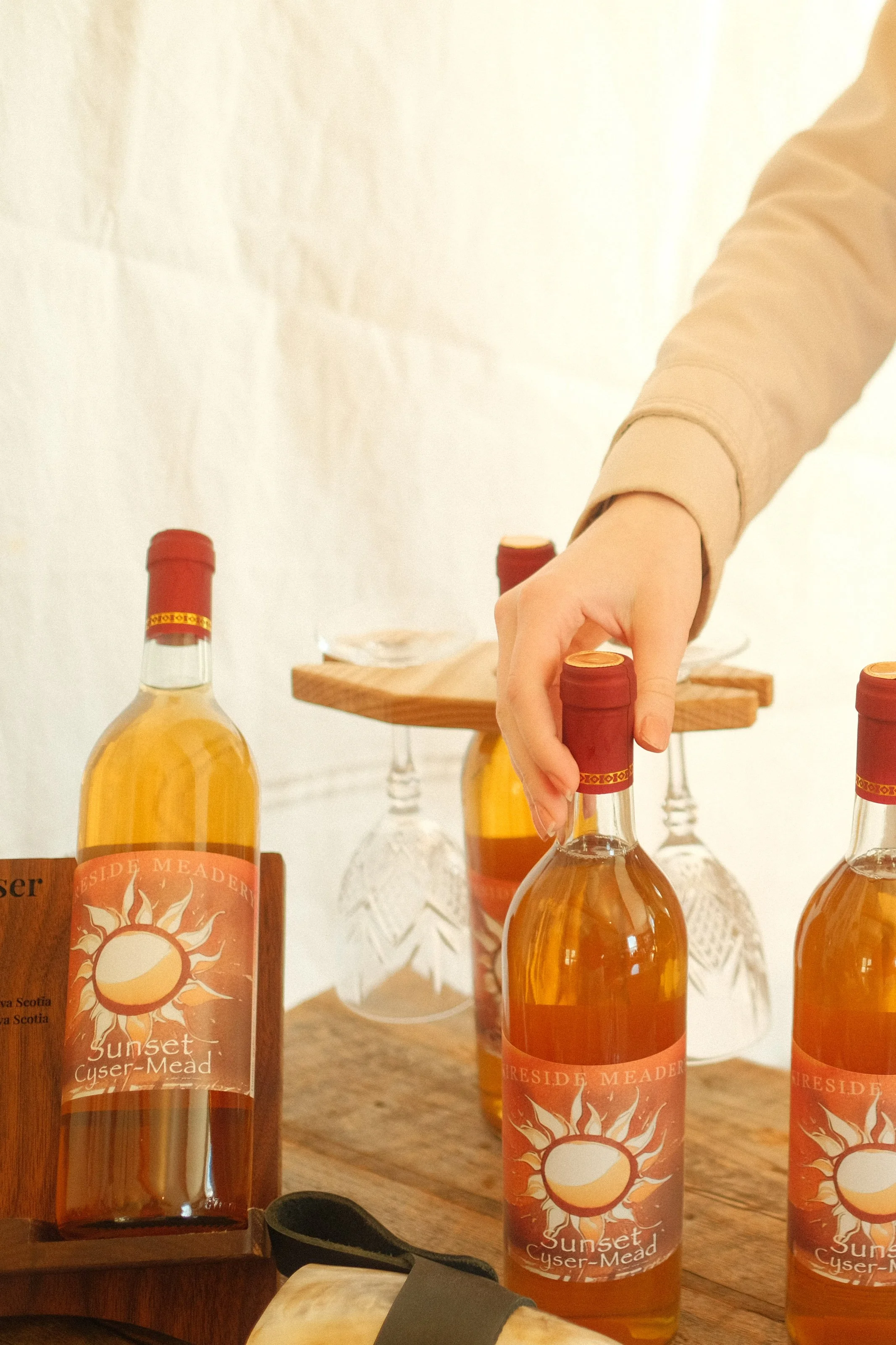 Person reaching for a bottle of Sunset Cysar-Mead on a wooden table, with more bottles and glasses of mead in the background.