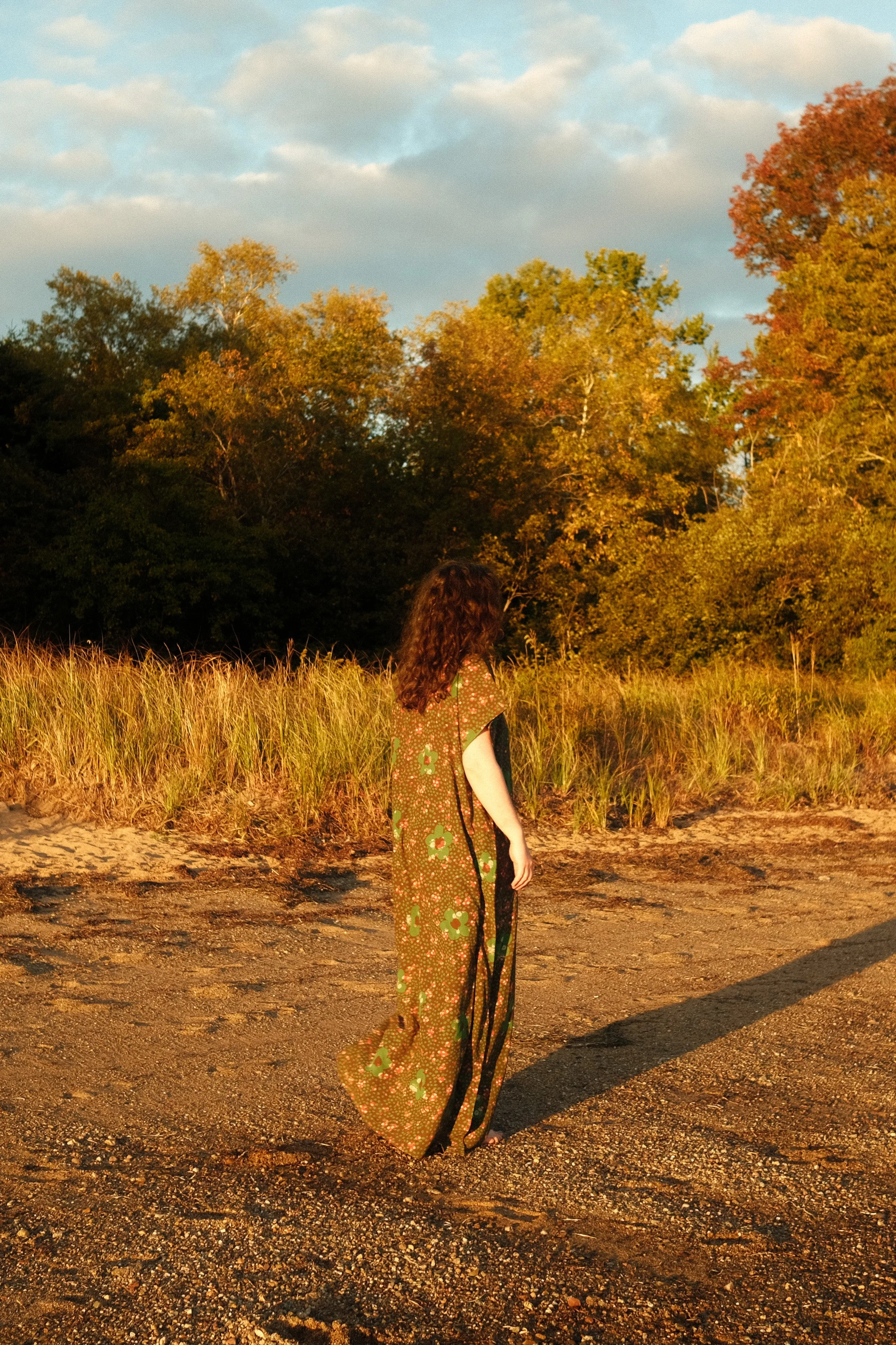 A woman with long, curly brown hair standing on a dirt path during sunset, with tall grass and colorful trees in the background.