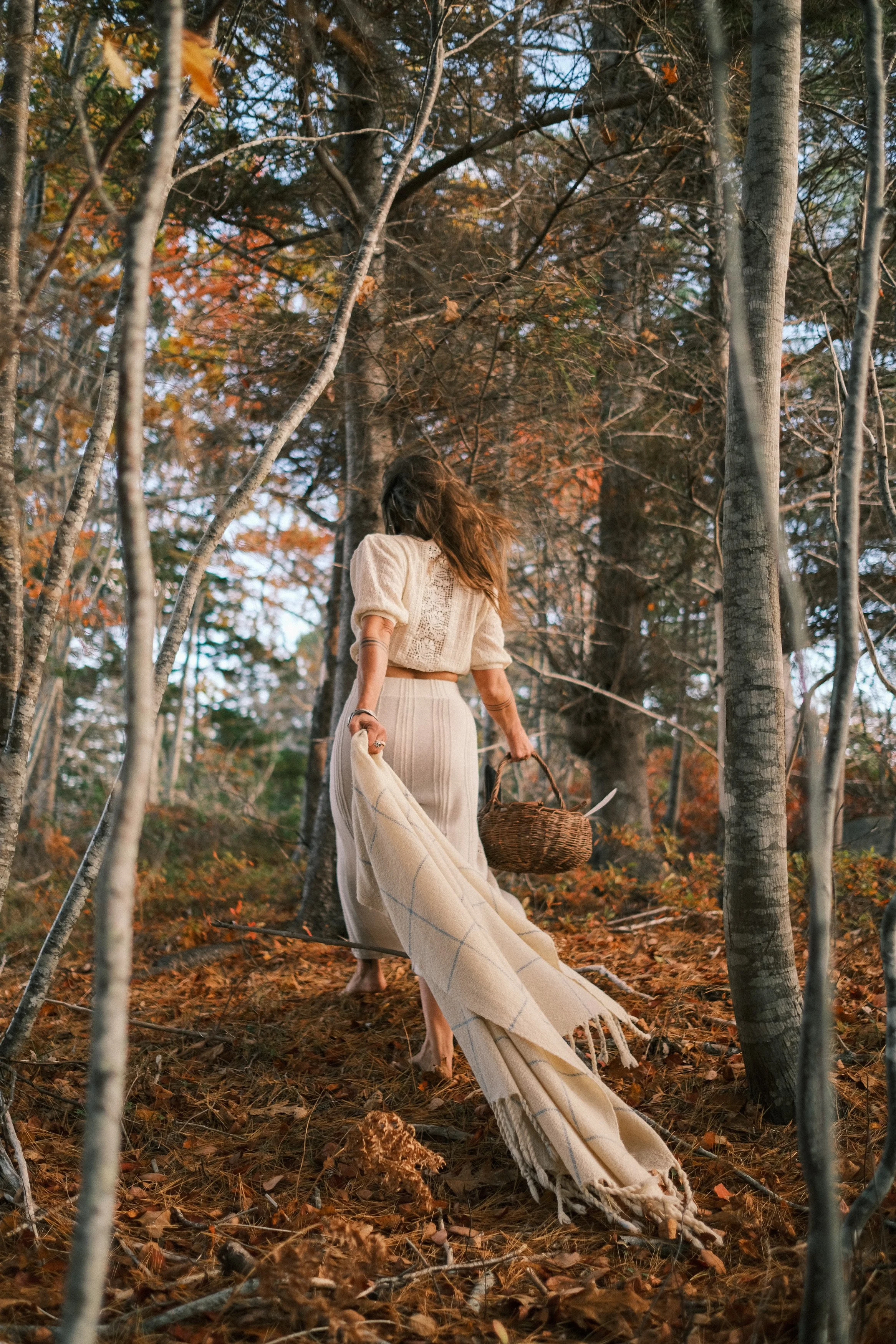 A woman is walking through a forest with autumn leaves on the ground, holding a woven basket and a beige blanket.