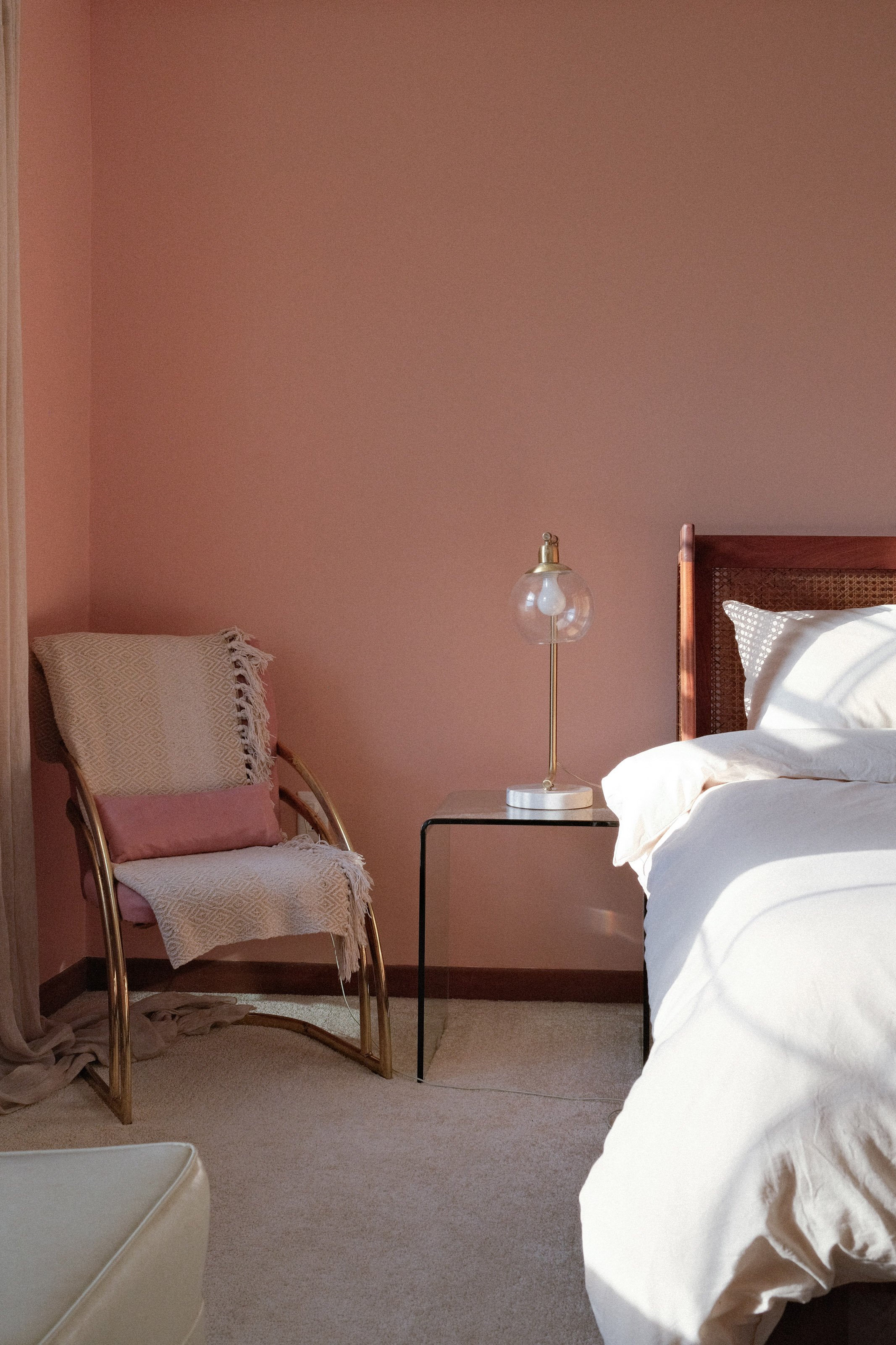 A bedroom with a pink wall, a bed with white bedding, a nightstand with a lamp, and a chair with pink and beige pillows and a blanket.