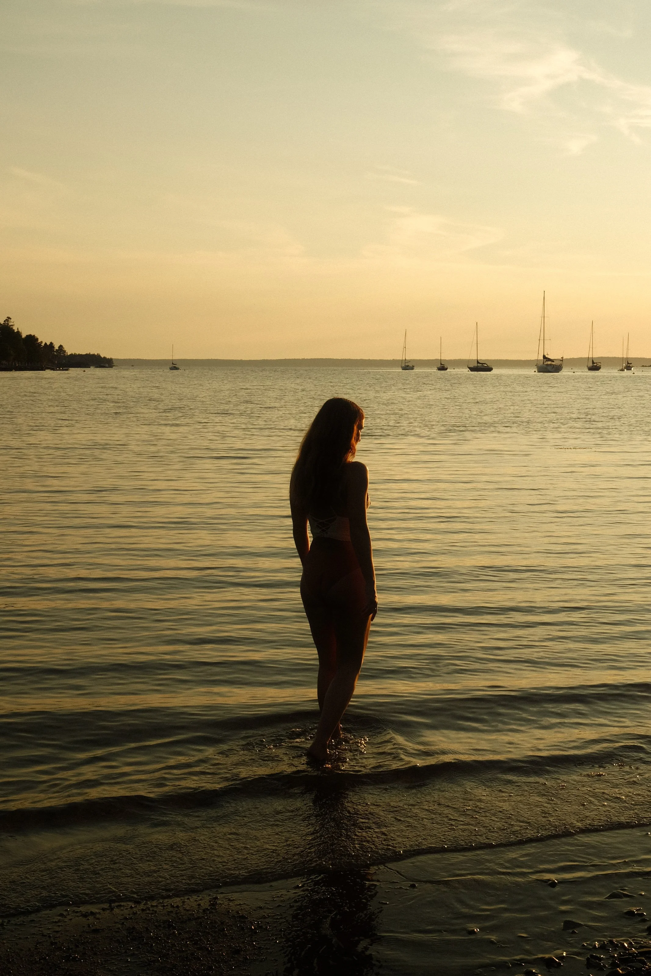 A woman standing in shallow water near a beach during sunset with sailboats in the background.