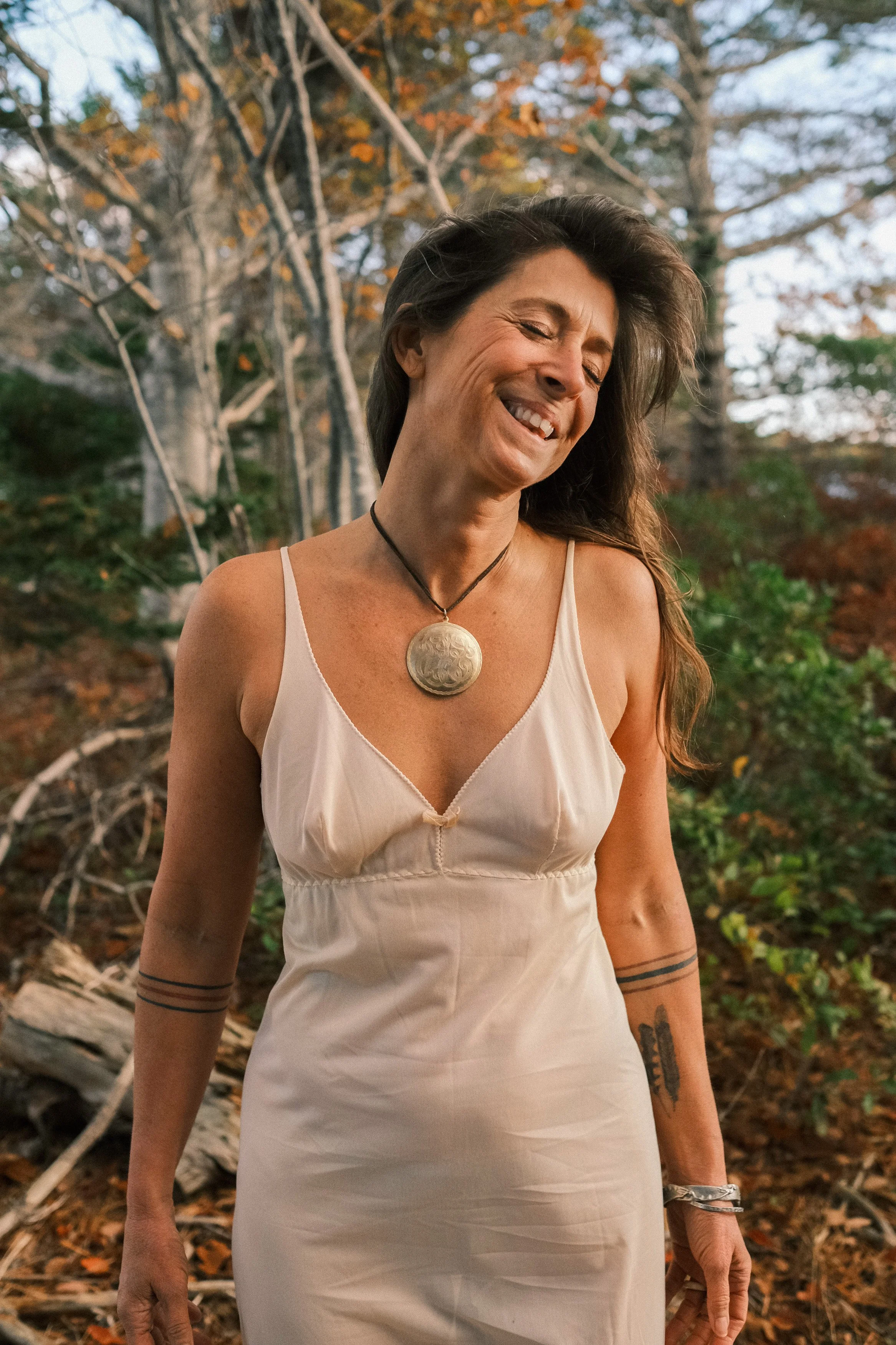 A woman with long dark hair smiling and wearing a white dress and a large metallic necklace standing outdoors with trees and autumn foliage in the background.
