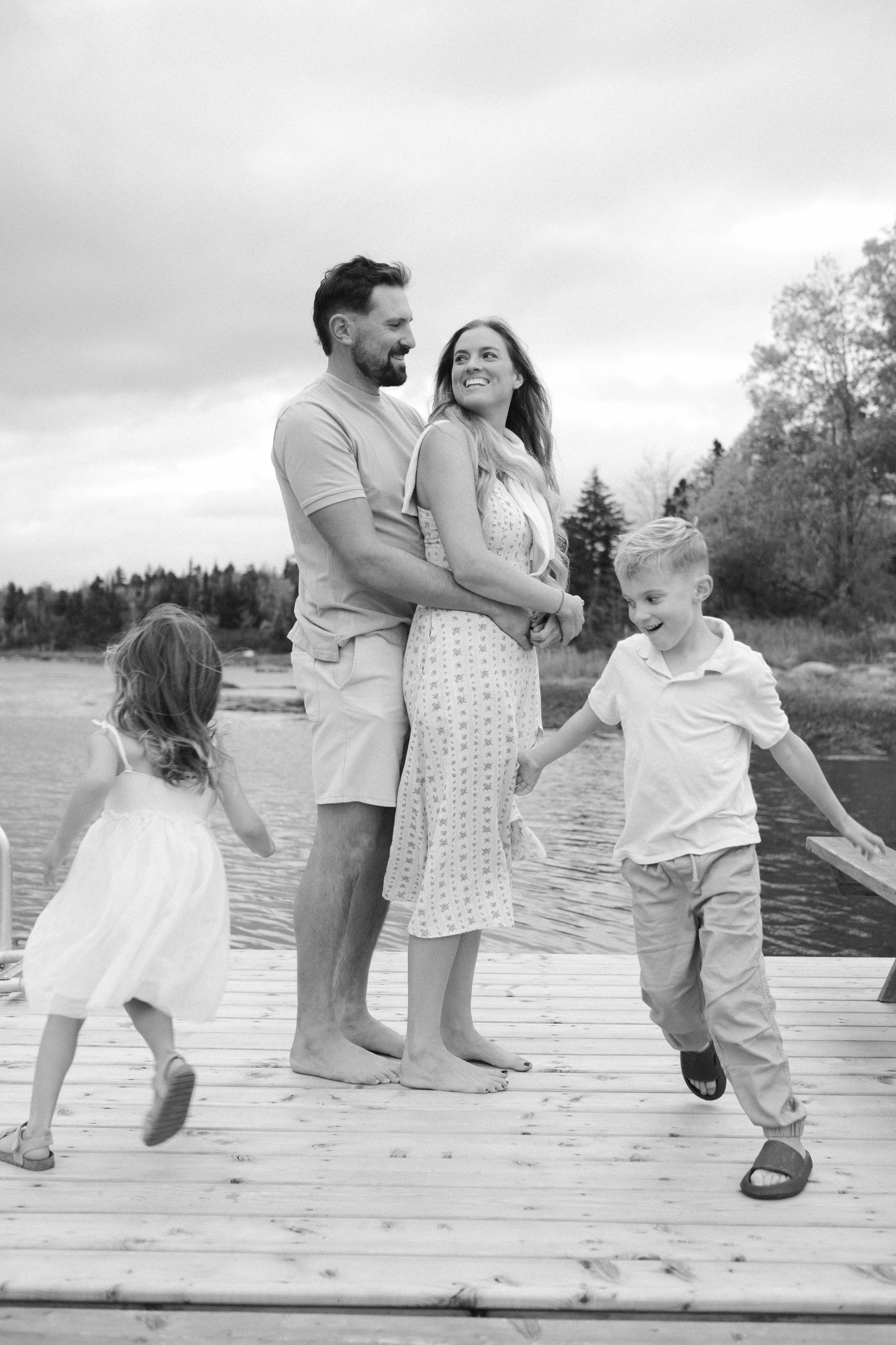 A happy family of four enjoying time together on a dock by a lake. The mother and father are standing close, smiling, with the children playing around them.