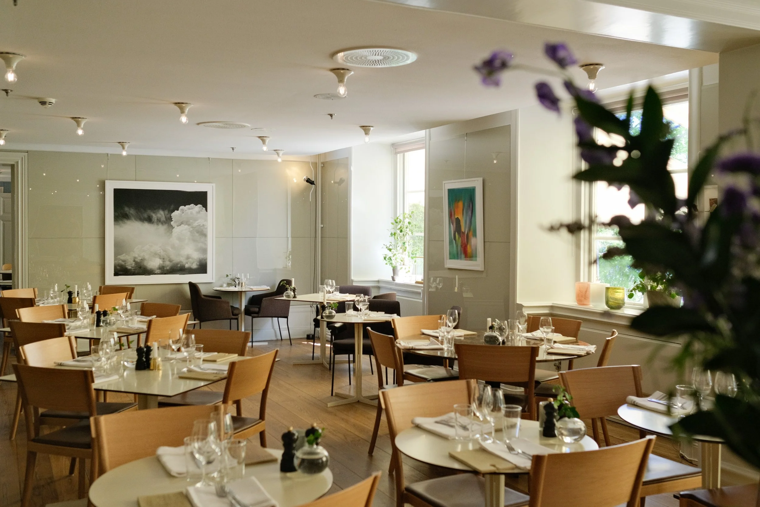 Empty restaurant dining area with wooden tables and chairs, set with glassware, white napkins, and small decorations, illuminated by natural light through window, with framed abstract artwork on walls.