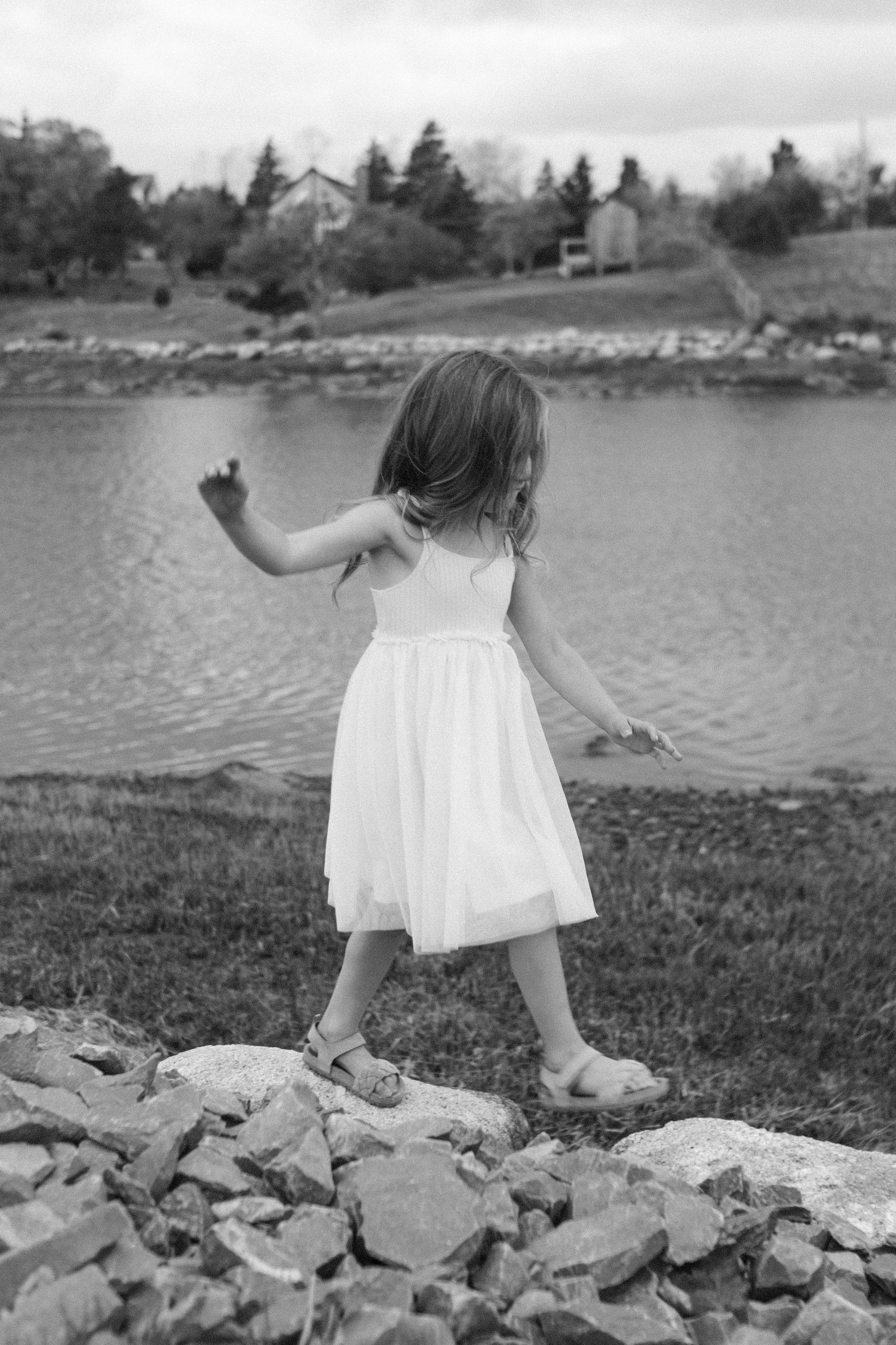 A young girl in a white dress and sandals walking on rocks near a body of water.