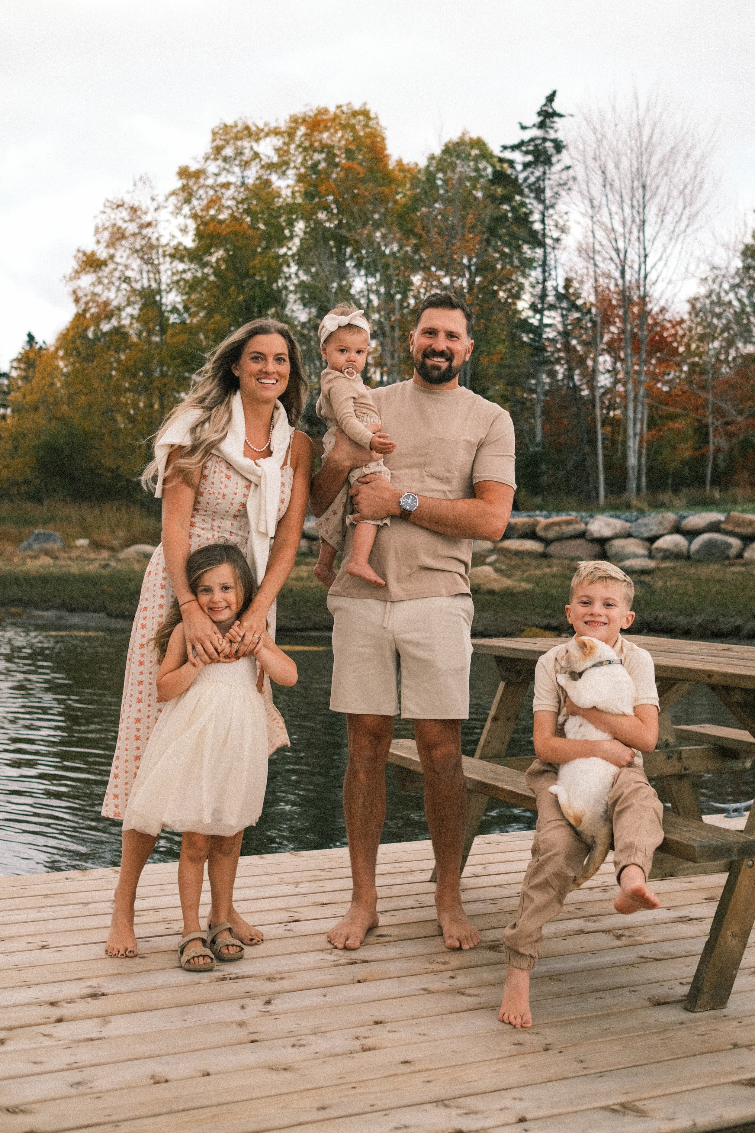 A family of six posing on a wooden dock near a lake with autumn trees in the background. The family includes two adults and four children, one of whom is a baby. They are smiling and appear happy.
