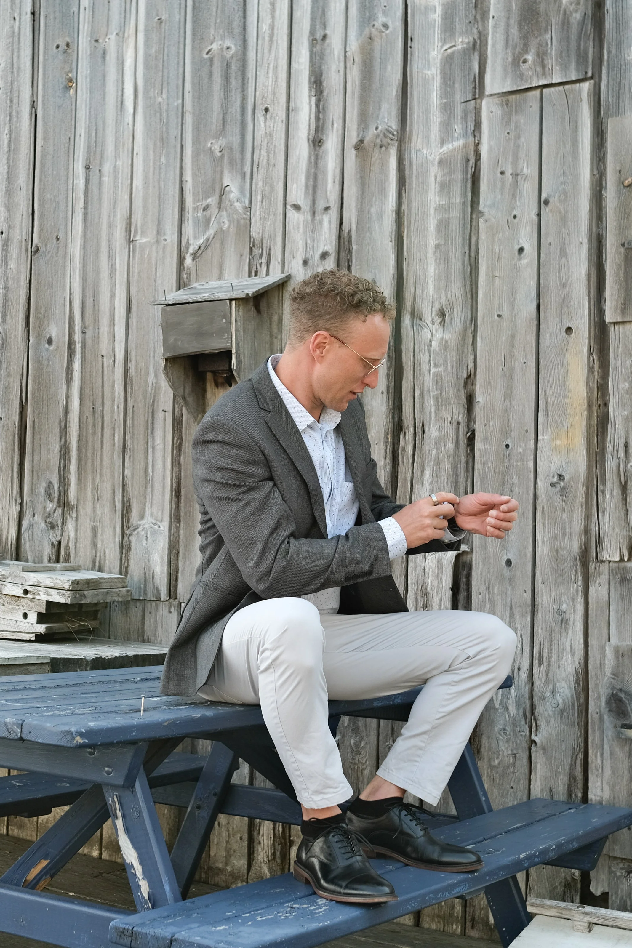 Man in gray blazer and white pants sitting on a blue picnic table, looking at his wristwatch, against a weathered wooden fence background.