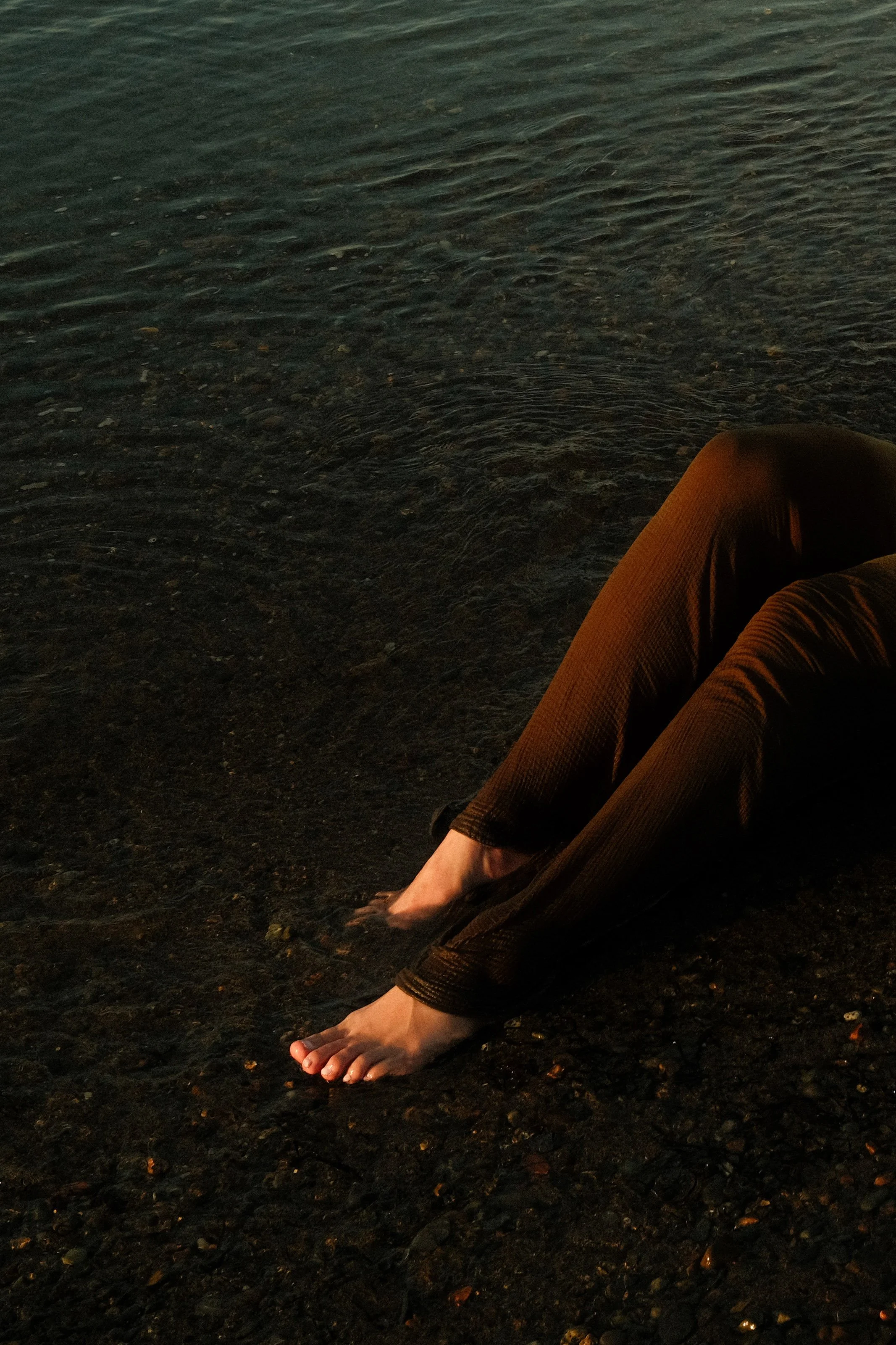 Person with brown pants sitting on a rocky shore with feet in the water, near a body of water at sunset or sunrise.